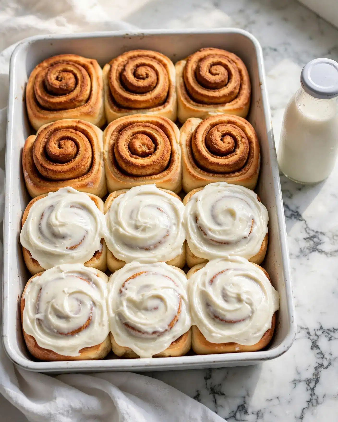 Soft and Fluffy Homemade Cinnamon Rolls Recipe 5 A white rectangular baking dish holds twelve cinnamon rolls arranged in four rows of three. The bottom eight rolls are thickly covered with smooth, creamy white frosting, showing soft swirls of cinnamon inside. The top four rolls are plain with a golden-brown baked crust and spiraled cinnamon layers, slightly rising above the others. The dish is placed on a white marbled surface with a white cloth partially underneath it, and a small white milk bottle is seen nearby. Photo taken with an iphone --ar 4:5 --v 7