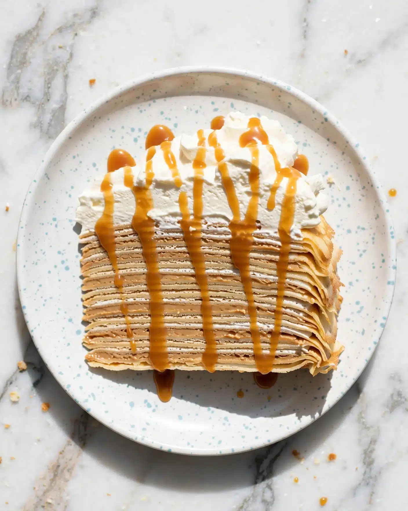 A slice of layered crepe cake with about 15 thin crepe layers separated by creamy light brown filling, topped with a thick white whipped cream layer drizzled with golden caramel sauce, placed on a white plate with light blue speckles, all on a white marbled surface. photo taken with an iphone --ar 4:5 --v 7