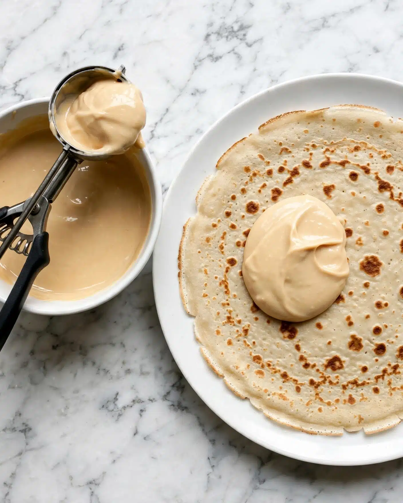 The image shows a white plate with a light beige crepe that has a smooth texture with some darker spots, lying flat on a white marbled surface. On top of the crepe, near the center, there is a dollop of creamy, pale caramel-colored sauce with a thick texture. To the left of the plate, there is a small white bowl filled with the same creamy sauce, slightly shiny and smooth. Resting on the edge of the bowl is a metal ice cream scoop with black handles, holding some of the sauce. A woman's hand is not visible in the image. The background is a white marbled texture. Photo taken with an iphone --ar 4:5 --v 7
