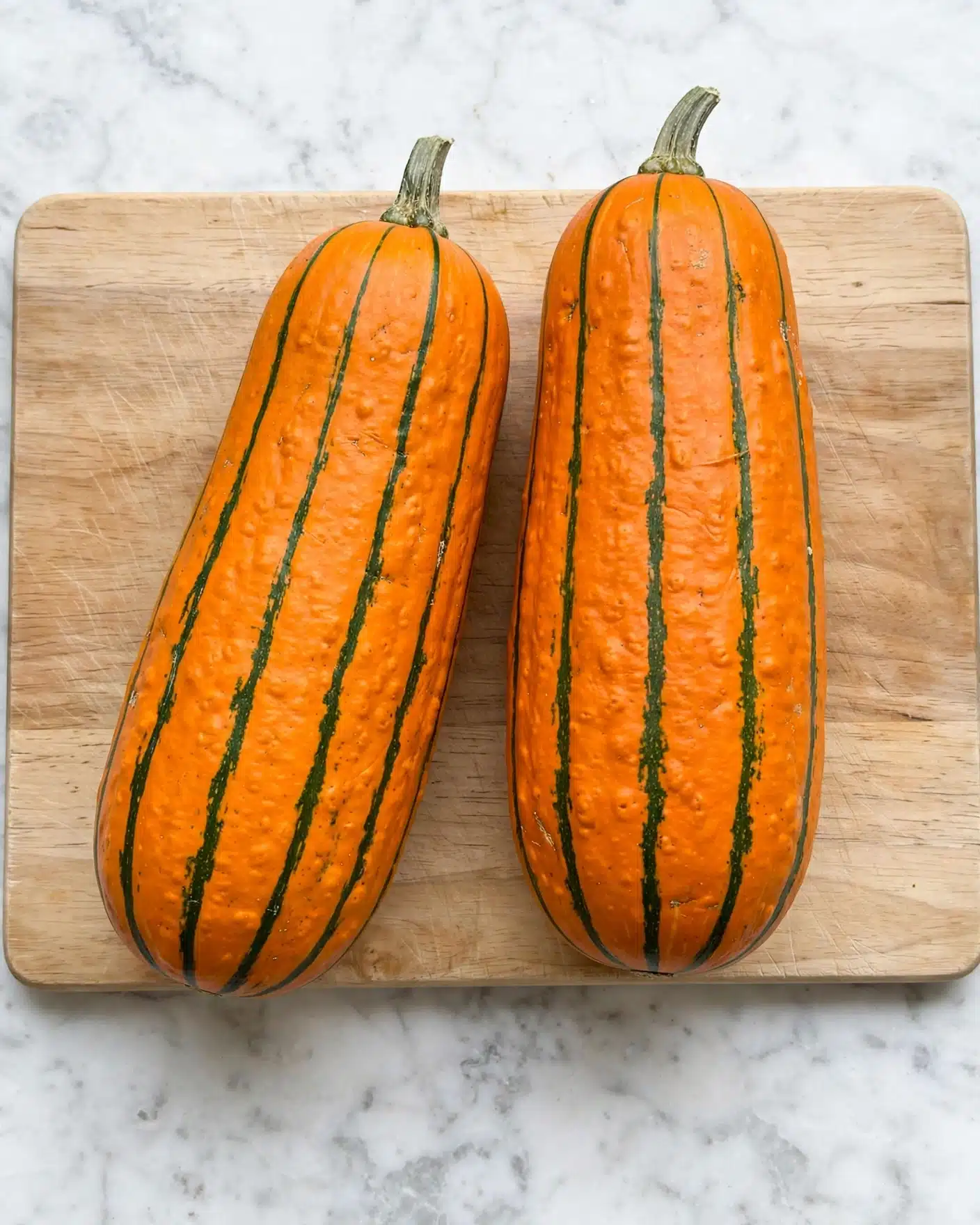 The image shows two whole orange and green striped squash placed side by side on a wooden cutting board with a light natural wood texture. The squash are elongated with rounded ends and have thin, vertical dark green stripes running from top to bottom on their orange skin. Each squash has a short greenish-gray stem at the top. The cutting board lies flat on a white marbled surface. The lighting is even, highlighting the colors and natural texture of the squash and board. photo taken with an iphone --ar 4:5 --v 7