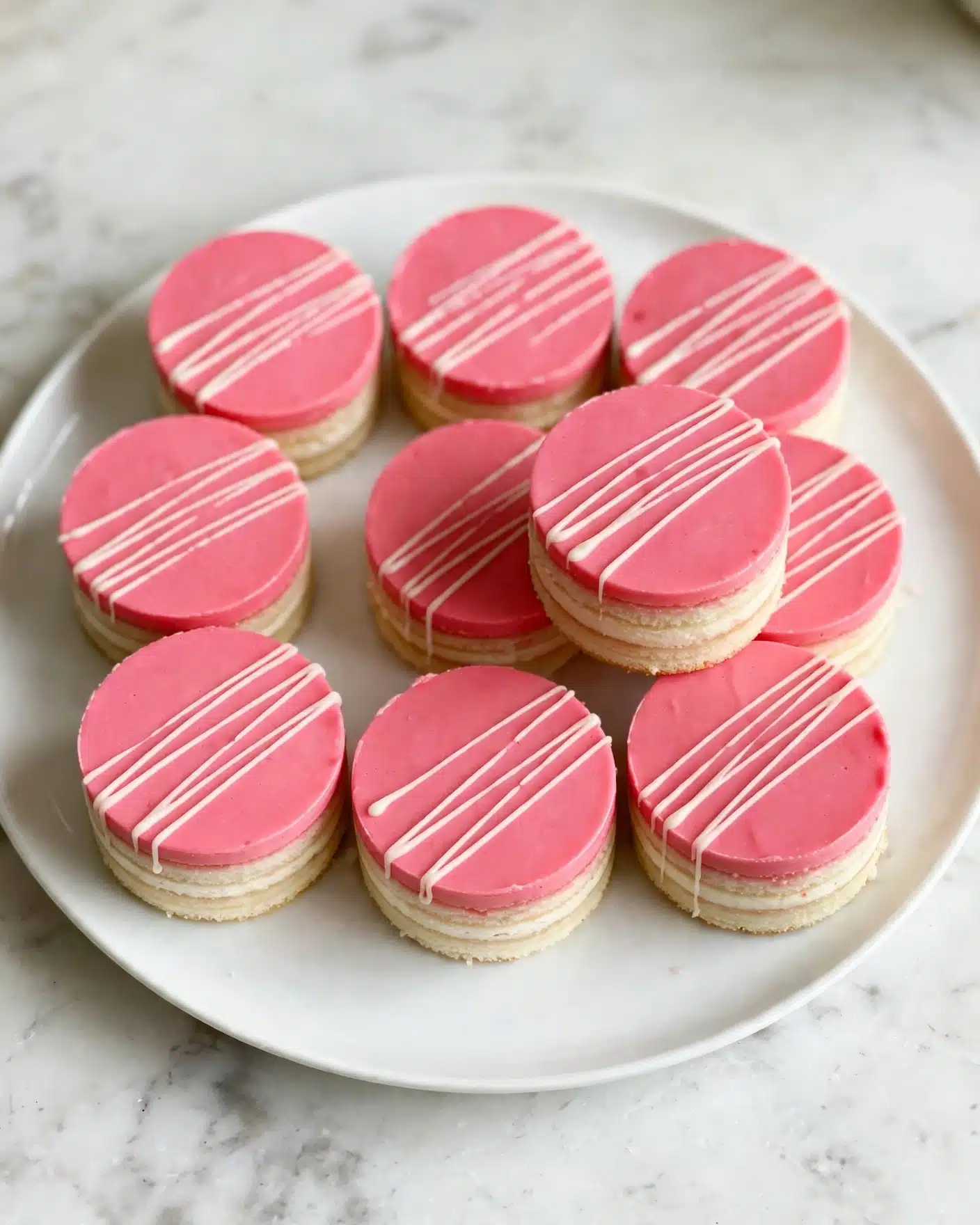 Strawberry Cake Pops Recipe 6 A white round plate holds nine small round desserts, each with a smooth, bright pink top layer. On top of each pink layer, there is a thin white drizzle pattern going across diagonally. The desserts have neat, straight sides with a hint of a lighter layer just visible at the base, suggesting multiple layers inside. One dessert is slightly stacked on another, showing their height and round shape clearly. The plate sits on a white marbled surface. photo taken with an iphone --ar 4:5 --v 7