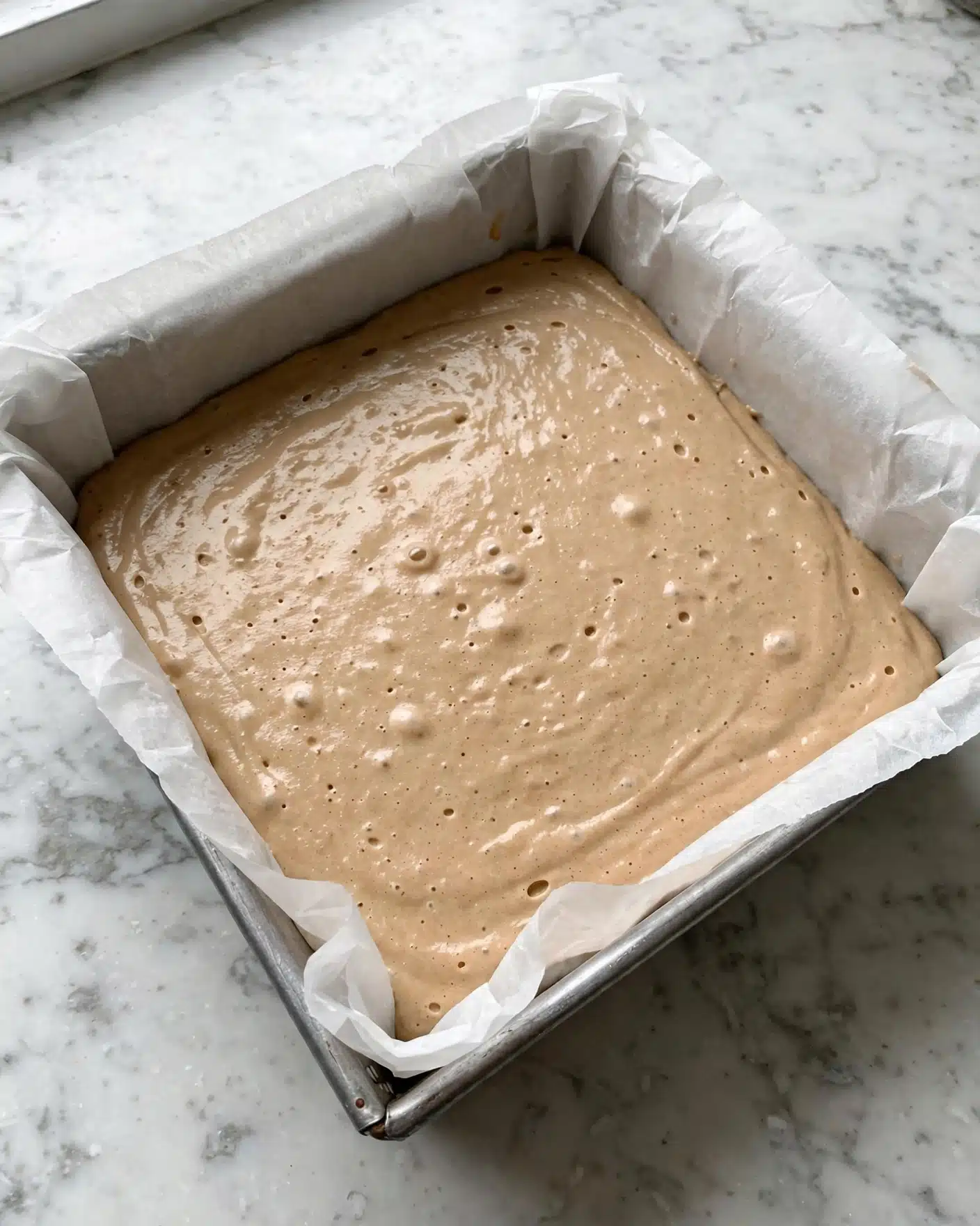 A square metal pan lined with white parchment paper holds an even layer of smooth, light brown batter spread flat inside. The batter has a creamy texture with a few small air bubbles on the surface. The pan sits on a white marbled surface. Photo taken with an iphone --ar 4:5 --v 7