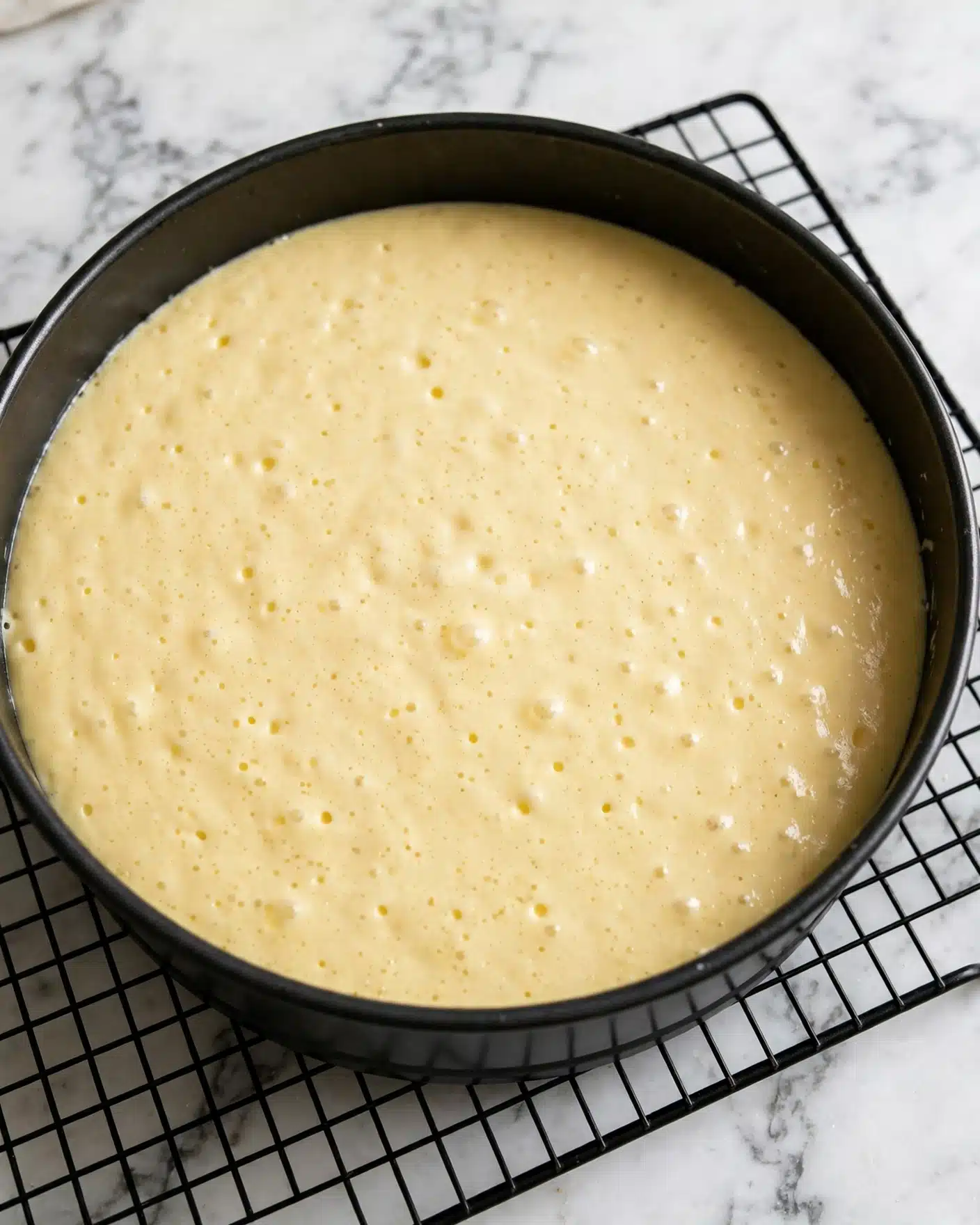 A round black baking pan filled with a smooth light yellow cake batter, placed on a black metal cooling rack. The batter surface is even and creamy with tiny bubbles visible. The background is a white marbled texture. photo taken with an iphone --ar 4:5 --v 7