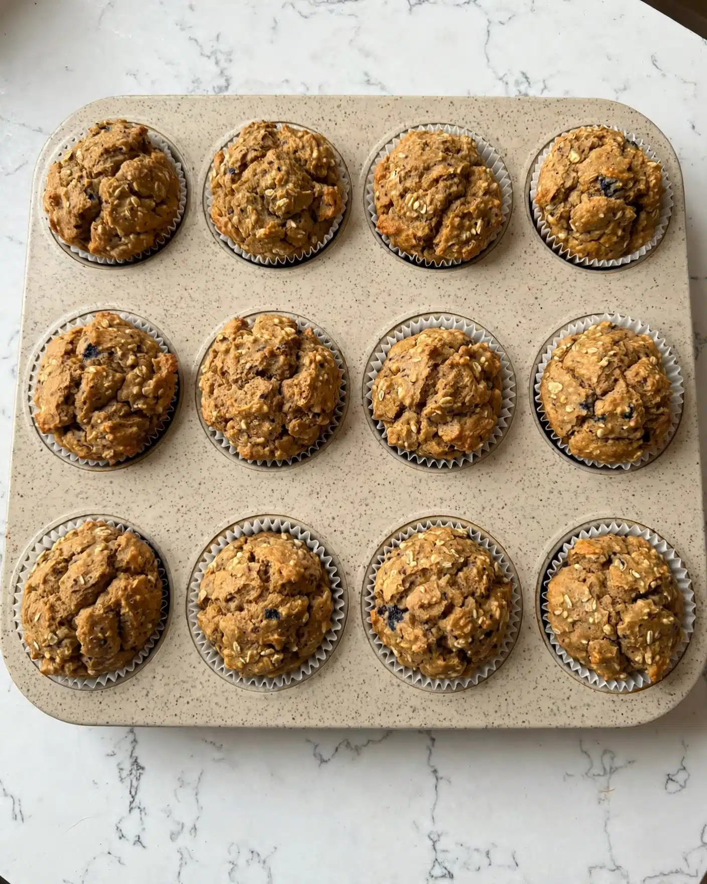 A beige muffin tray with a speckled pattern, holding 12 freshly baked muffins lined up in three rows of four. Each muffin has a golden-brown rough top with cracks and a slightly uneven texture, showing tiny bits of dark and light specks throughout. The paper liners inside each muffin cup are white with a ruffled edge. The tray sits on a white marbled surface with delicate gray veins. photo taken with an iphone --ar 4:5 --v 7