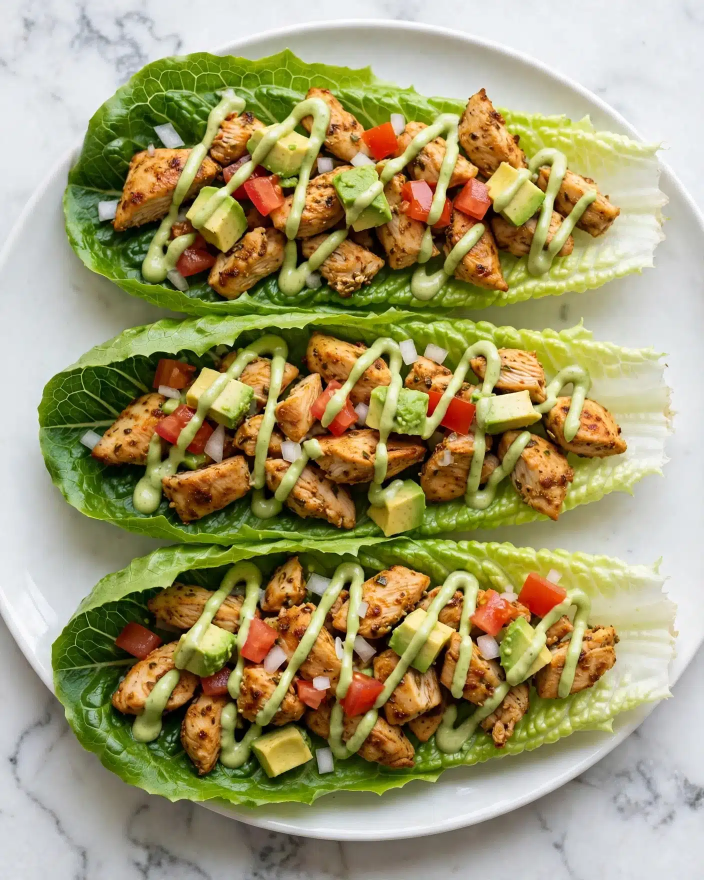 Three lettuce wraps are placed on a white plate with a white marbled background. Each wrap has three main layers: the dark green leafy lettuce at the bottom, topped with small chunks of cooked brown chicken, diced red tomatoes, and small cubes of light green avocado. A green sauce is drizzled over the top, covering parts of the chicken and avocado pieces. The textures range from soft avocado to slightly crispy chicken and fresh lettuce leaves with visible veins. The image is close up, showing the fresh colors and details. photo taken with an iphone --ar 4:5 --v 7