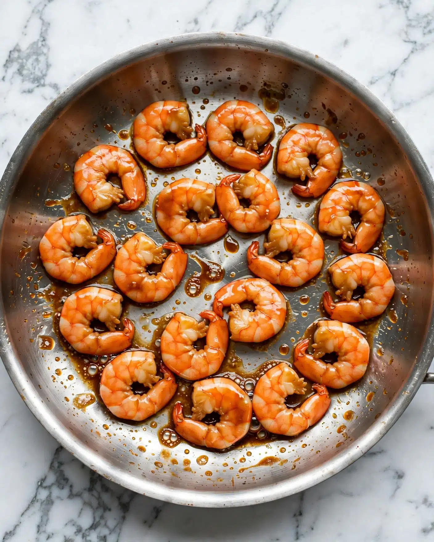 A round silver pan filled with 14 cooked shrimp arranged in a loose circular pattern, each shrimp plump and bright orange with a light glaze of shiny brown sauce coating them evenly, small bubbles and sauce stains visible around the shrimp on the pan’s surface, all set on a white marbled background photo taken with an iphone --ar 4:5 --v 7
