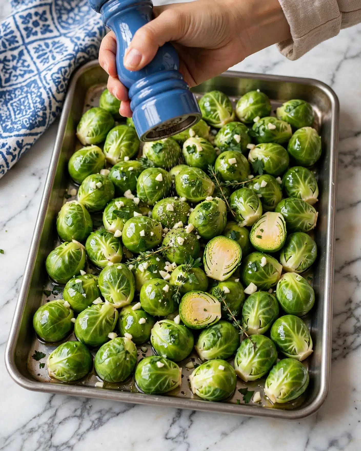 The image shows a metal baking tray filled with fresh Brussels sprouts that are bright green and shiny with olive oil. On top of the sprouts, there are small white chopped garlic pieces and some scattered fresh green herbs. A woman's hand is holding a blue pepper grinder above the tray, adding seasoning. The tray is placed on a white marbled surface, and a blue and white patterned cloth is seen at the top left corner. The overall scene looks fresh and ready to be cooked. photo taken with an iphone --ar 4:5 --v 7