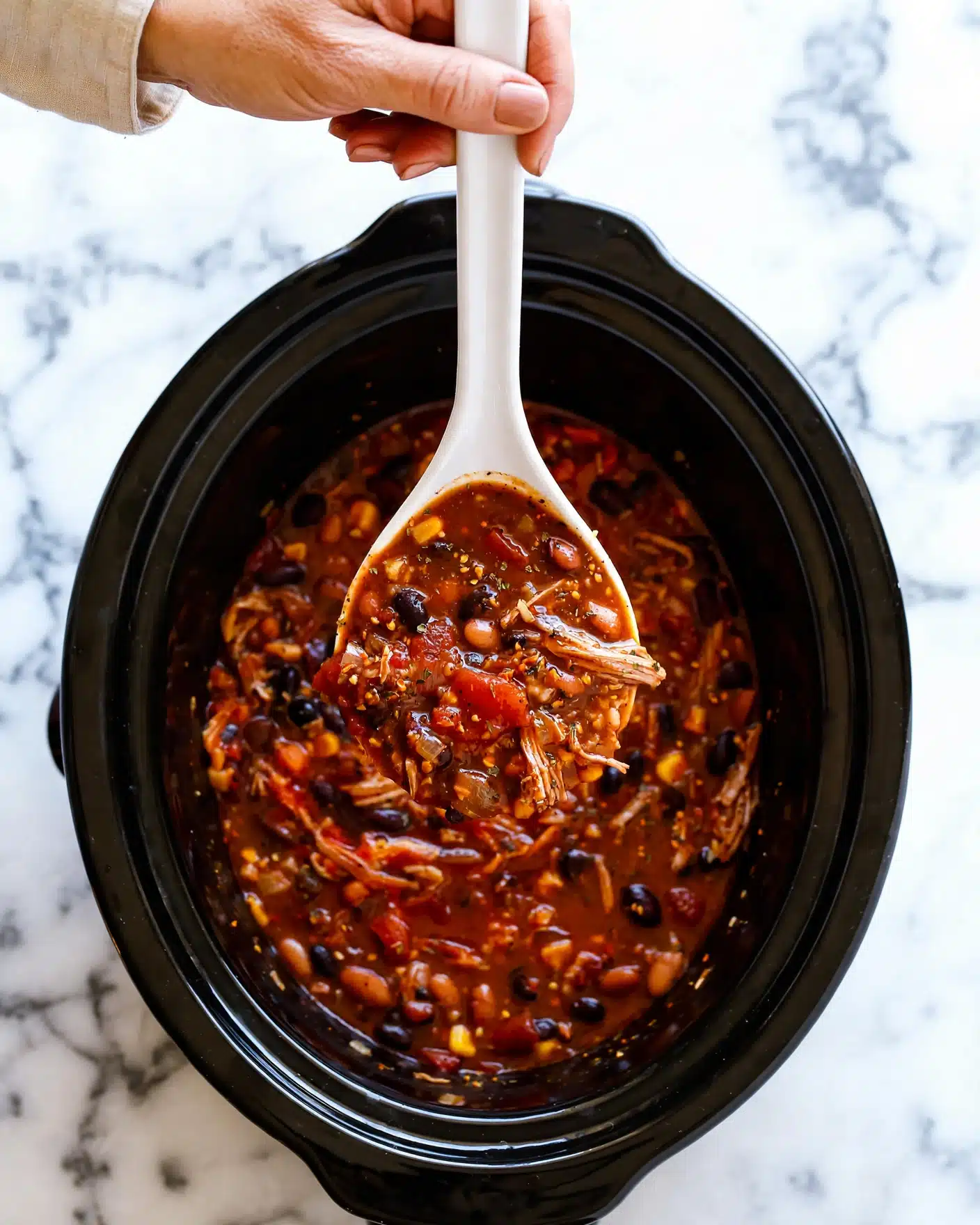 A white ladle lifting a thick, rich stew from a black slow cooker. The stew has many layers, starting with a deep reddish-brown sauce filled with small beans, grains, and shredded meat pieces, mixed together with visible chunks of tomato and spices. The texture looks hearty and chunky with a glossy surface. The black slow cooker contrasts with the white marbled background, and a woman's hand holds the ladle from above. photo taken with an iphone --ar 4:5 --v 7