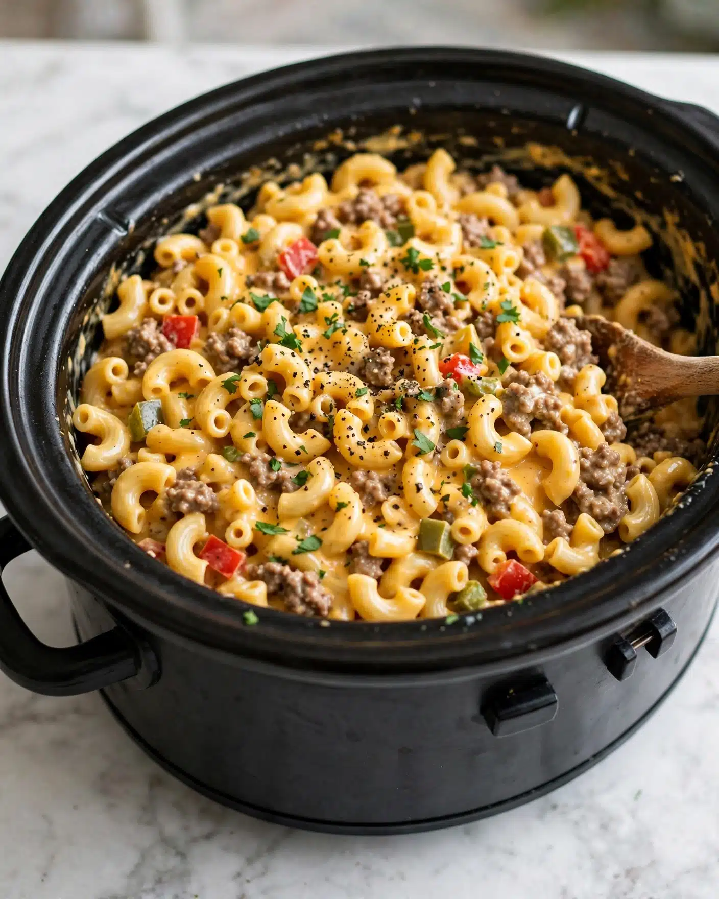 A black slow cooker filled with creamy macaroni pasta mixed with cooked ground meat and small pieces of red and green bell peppers, all coated in a smooth, cheesy sauce. The pasta is elbow-shaped and yellow-orange in color, and the dish is garnished with chopped green herbs and a sprinkling of black pepper on top. A wooden spoon is on the right side, partially submerged in the macaroni and meat mix. The slow cooker is placed on a white marbled surface. photo taken with an iphone --ar 4:5 --v 7