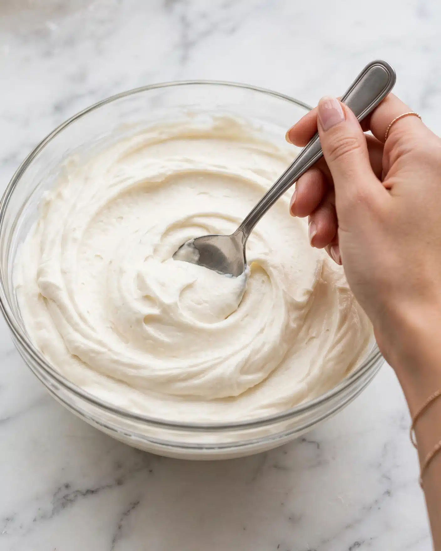 A clear glass bowl filled with a thick layer of smooth, creamy white frosting that has soft swirls and peaks on the surface, with a woman's hand holding a metal spoon gently resting in the frosting, all set on a white marbled surface. Photo taken with an iphone --ar 4:5 --v 7