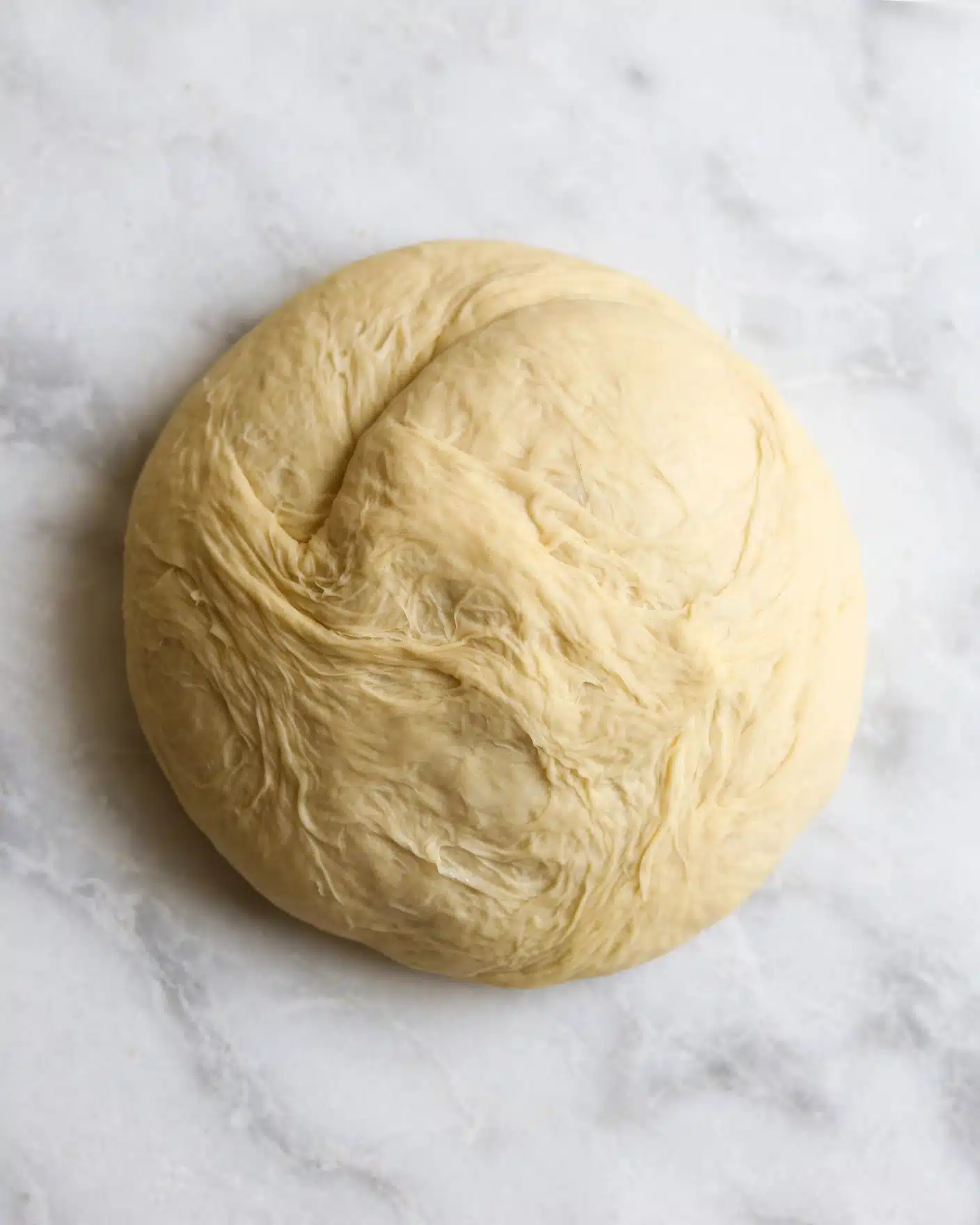 A single round dough ball with a smooth, slightly uneven surface rests on a white marbled texture. The dough has a light yellow-beige color with a soft, matte finish, showing faint textures of stretching and folding throughout. The edges are softly rounded, blending gently into the center, which appears slightly thicker and more compact. Photo taken with an iphone --ar 4:5 --v 7