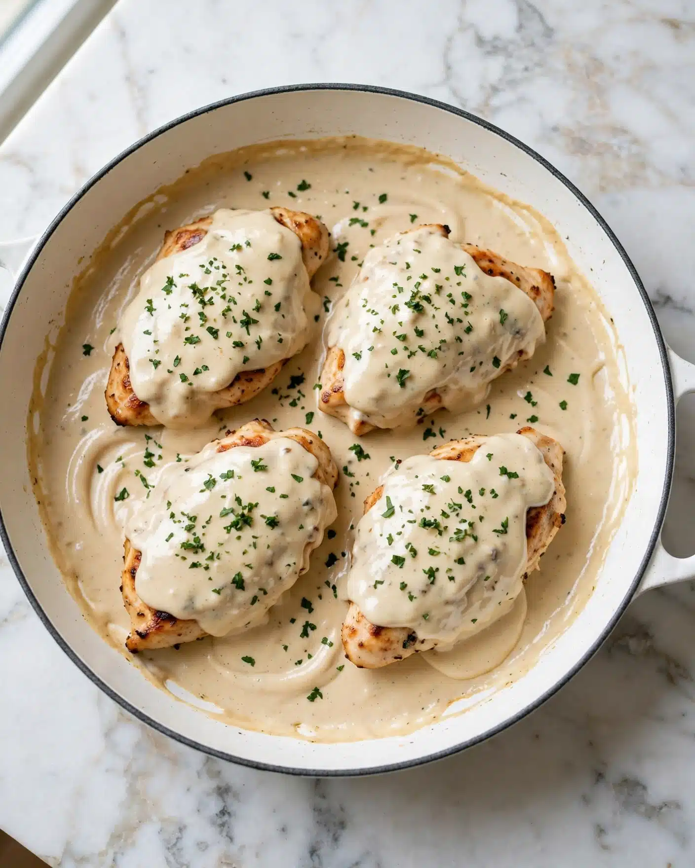 The image shows a dish with four pieces of golden-brown grilled chicken on the bottom layer, placed in a white skillet. The chicken is covered by a thick creamy light beige sauce with smooth texture, spread evenly over the chicken and the skillet base. Small bits of green herbs are sprinkled on top across the creamy sauce, adding color and texture contrast. The skillet is set on a white marbled surface. photo taken with an iphone --ar 4:5 --v 7