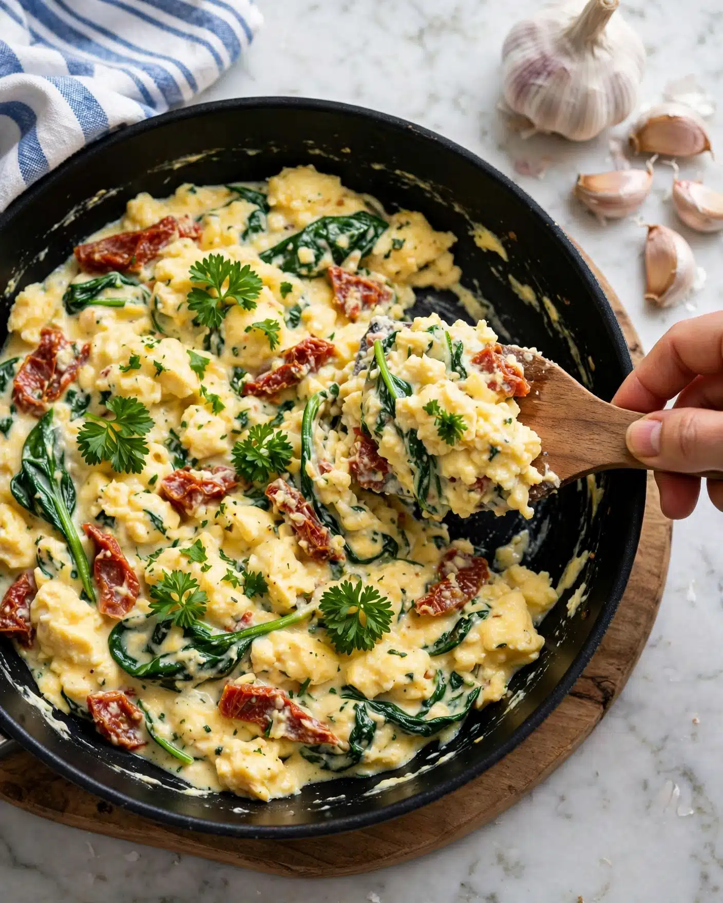Tuscan Spaghetti Squash in Creamy Sun-Dried Tomato and Spinach Sauce Recipe 6 A black pan filled with a creamy yellow scrambled egg mix that has green spinach leaves and red sun-dried tomato pieces spread throughout. The eggs have a soft, slightly lumpy texture with small sprigs of bright green parsley sprinkled on top. A woman's hand is holding a wooden spatula, lifting some of the eggs on the right side of the pan. The pan is resting on a wooden board placed on a white marbled surface, with a whole garlic bulb and garlic cloves visible in the upper right corner. A blue and white striped cloth is positioned at the top left near the pan edge. Photo taken with an iphone --ar 4:5 --v 7