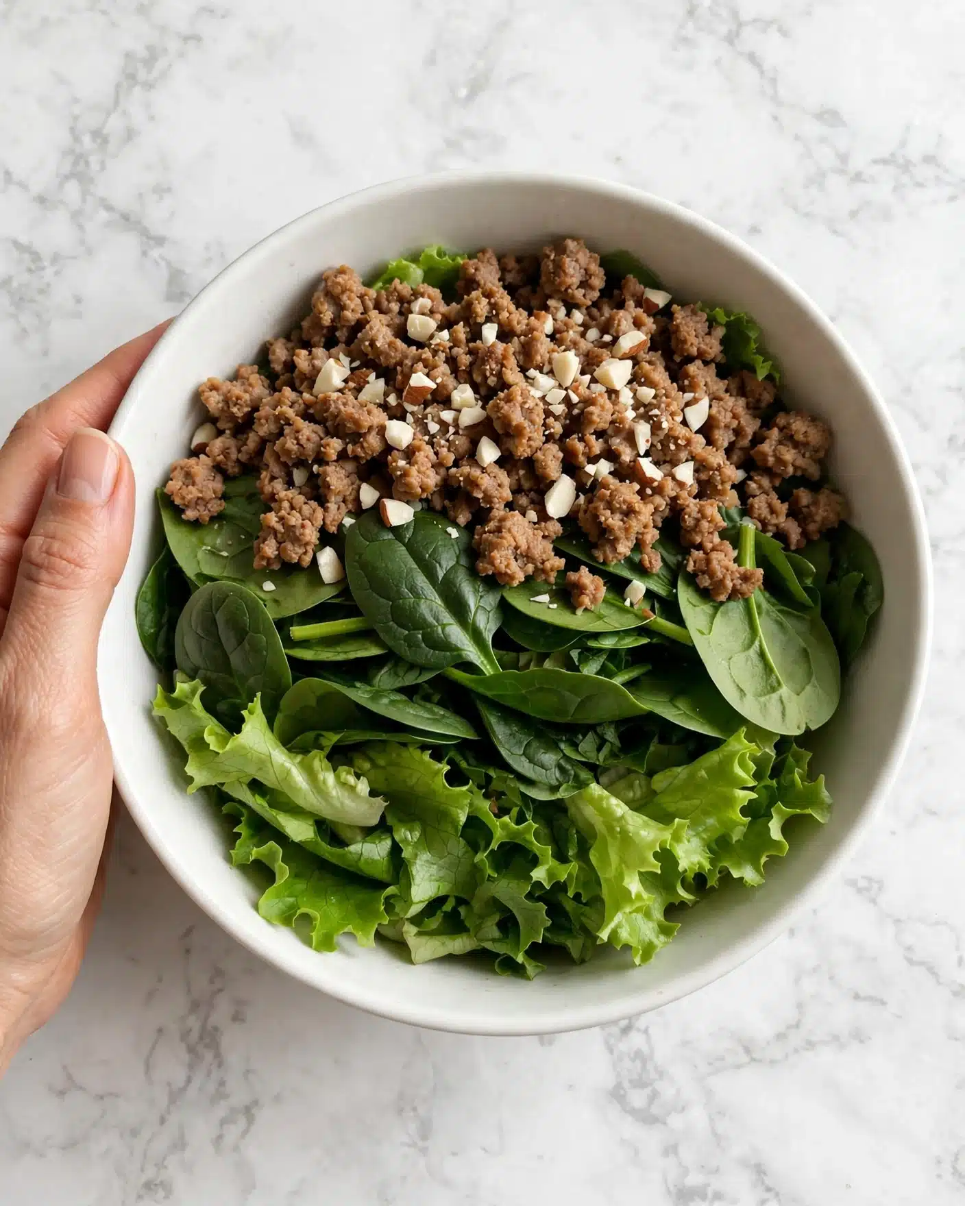 A white bowl filled with a fresh salad showing three main layers: the bottom layer is bright green leafy lettuce, the middle layer consists of dark green spinach leaves, and the top layer features cooked ground meat that is lightly browned with some small pieces of white chopped nuts sprinkled over it. The bowl sits on a white marbled surface, and a woman's hand is gently holding the bowl on the left side. photo taken with an iphone --ar 4:5 --v 7