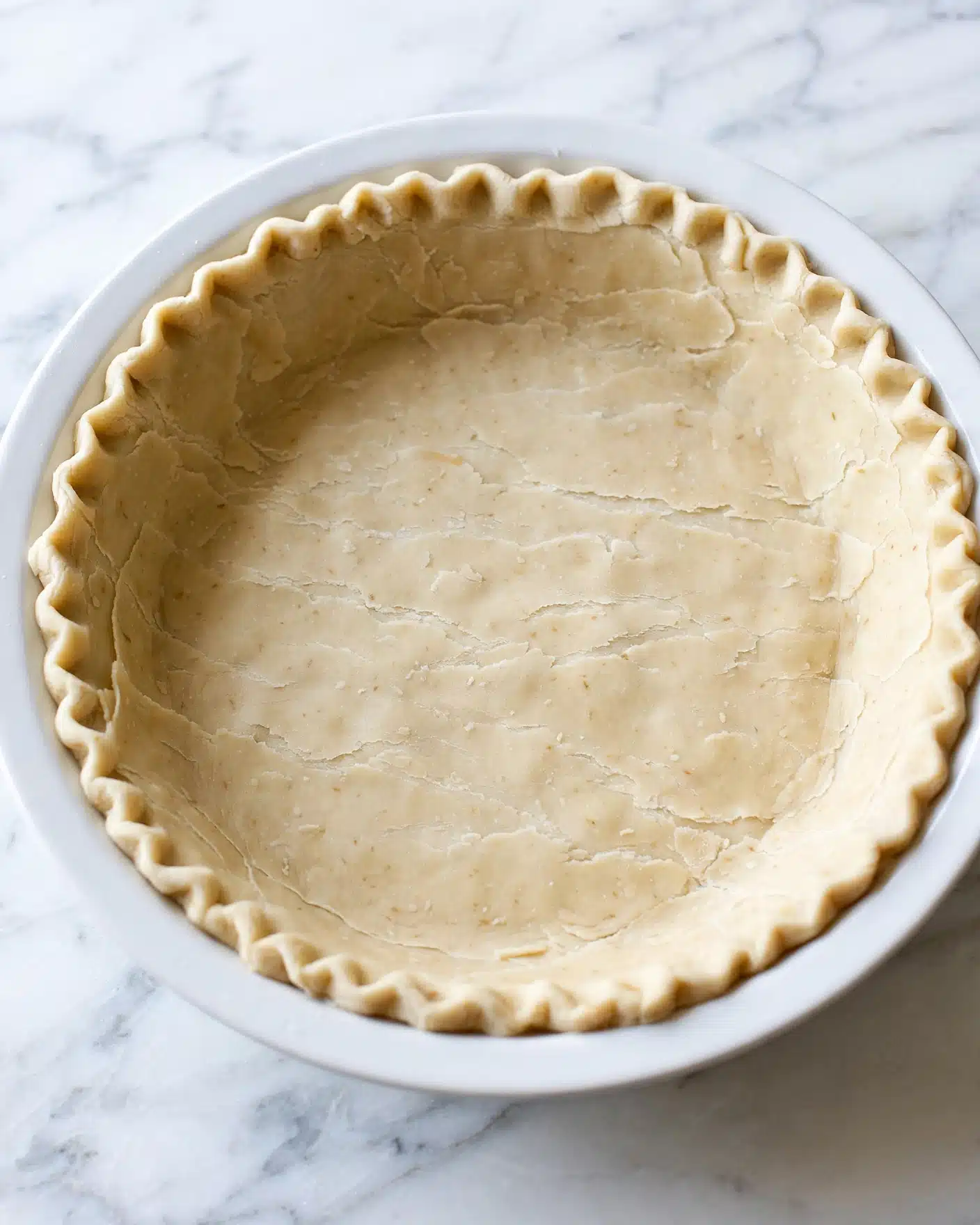 Walnut Pie Recipe 5 The image shows an uncooked pie crust filling a white pie dish. The crust is light beige with a smooth texture spread evenly across the dish. The edge is decorated with a simple, regular crimped pattern all around the top rim. The pie dish sits on a white marbled surface with soft lighting that highlights the dough’s slightly flaky texture. Photo taken with an iphone --ar 4:5 --v 7
