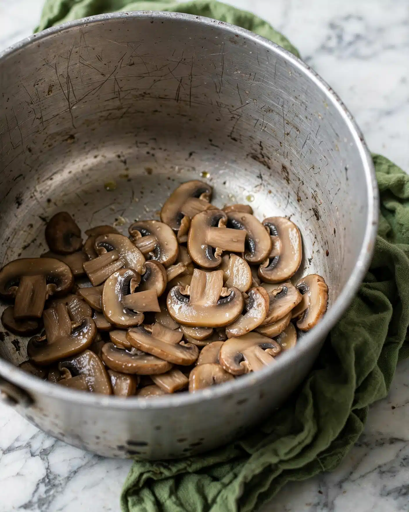 A close-up image of cooked, sliced mushrooms inside a silver metal pot. The mushrooms are light to medium brown in color with a slightly shiny and soft texture, and they sit loosely at the bottom layer of the pot. The pot has scratches and marks inside, and it rests on a crumpled green cloth on a white marbled surface background. The overall scene feels earthy and simple, focusing on the mushrooms and the pot photo taken with an iphone --ar 4:5 --v 7