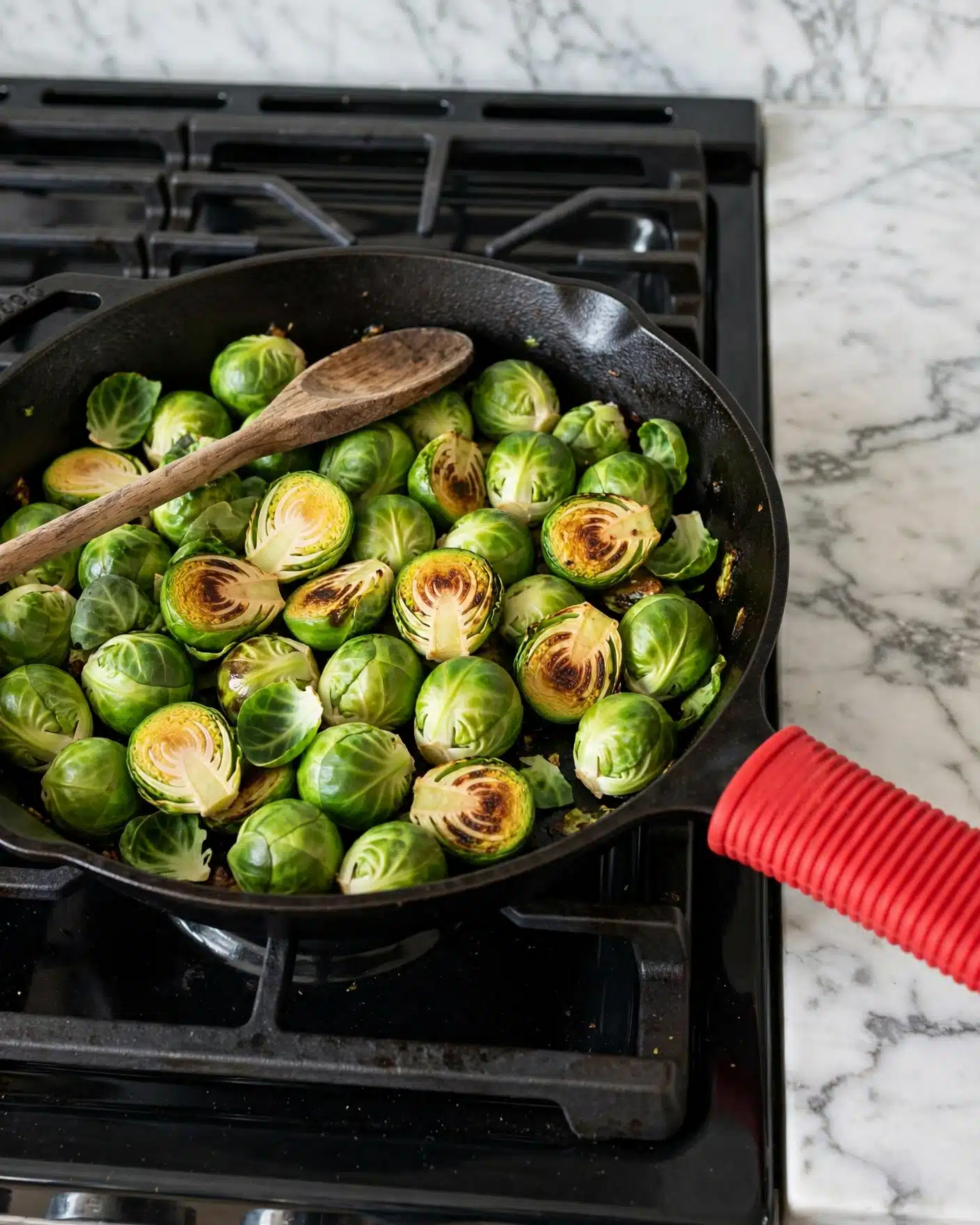 A black cast iron pan with a red handle edge filled with bright green Brussels sprouts, some whole and some sliced in half, showing charred, golden-brown spots on the cut sides. The Brussels sprouts have fresh green leaves mixed in, and there is a wooden spoon resting inside the pan on top of the sprouts. The pan is placed on a black stovetop with metal grates, and the surrounding surface is a white marbled texture. photo taken with an iphone --ar 4:5 --v 7