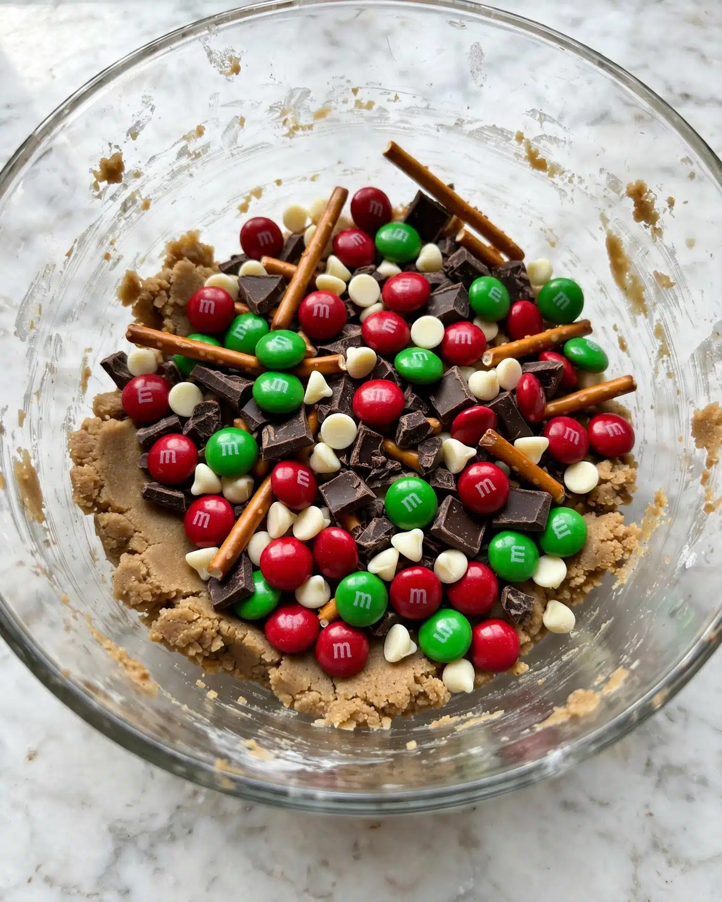 A clear glass bowl filled with a mix of colorful candy and snacks. The bottom layer is a light brown cookie dough with a rough texture. On top of this are scattered red and green chocolate candies with a smooth, shiny surface and a small 
