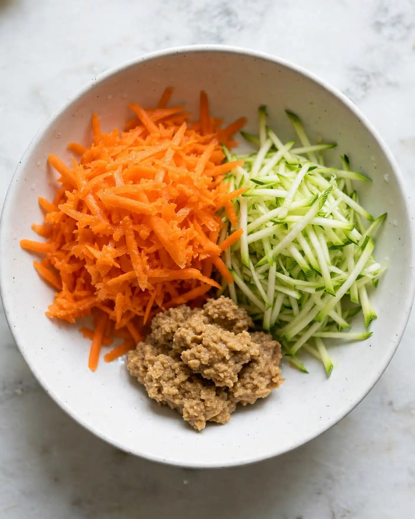 The image shows three main elements arranged on a white marbled surface: in the front, there is a small pile of shredded bright orange carrots with a slightly moist texture; behind and slightly to the right is a similar pile of finely shredded pale green zucchini; to the left, there is a small white bowl filled with a chunky light brown sauce or paste that looks thick and coarse. The overall appearance is fresh and simple. Photo taken with an iphone --ar 4:5 --v 7