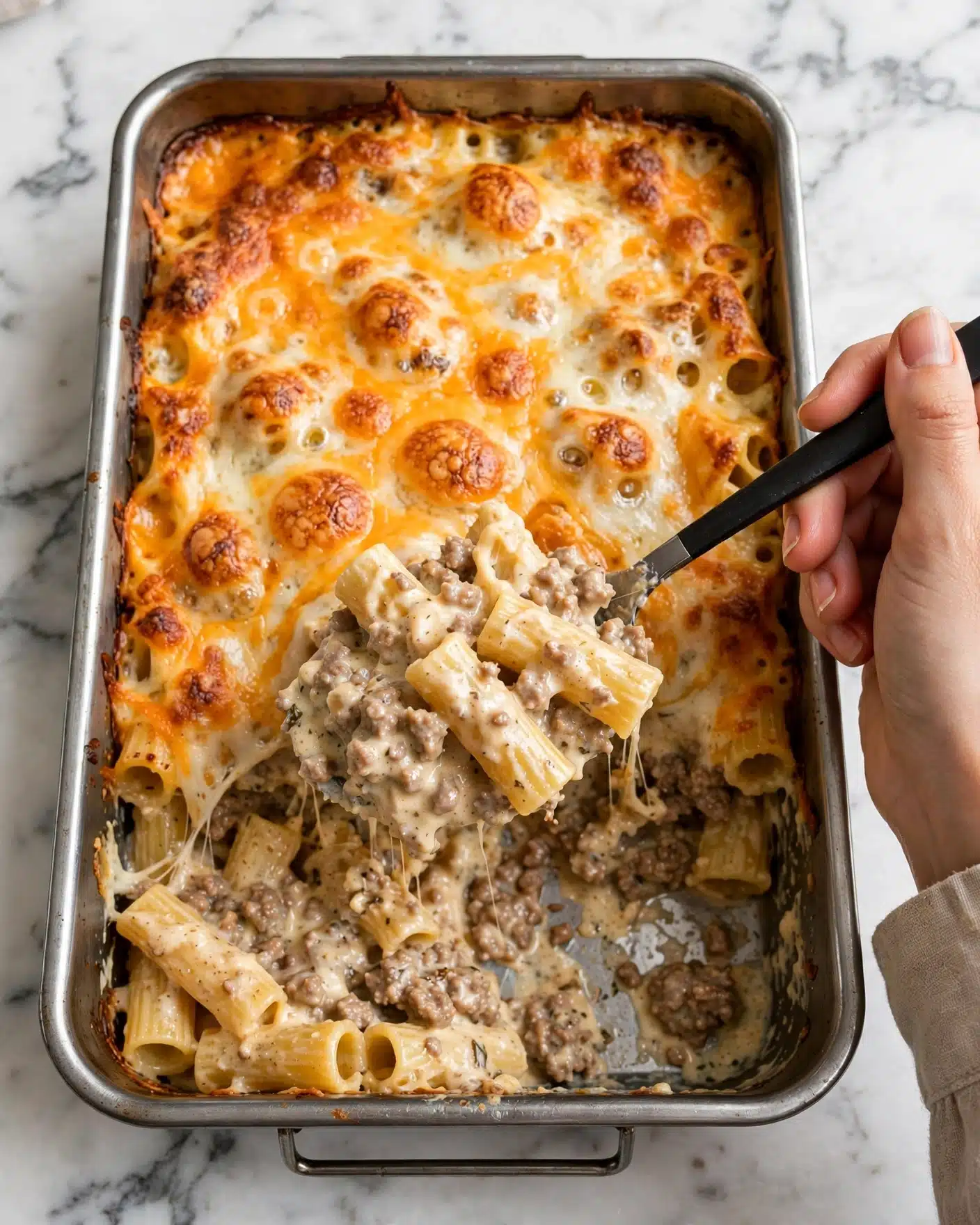 A rectangular metal tray filled with a baked dish showing a top layer of melted orange and white cheese that looks slightly browned and bubbly. The dish inside has a thick layer of pasta mixed with ground meat and sauce, visible as a spoon lifts a portion out. The pasta appears coated with a creamy sauce showing brown meat bits mixed evenly throughout. The background is a white marbled surface with a woman’s hand holding the black-handled spoon. Photo taken with an iphone --ar 4:5 --v 7