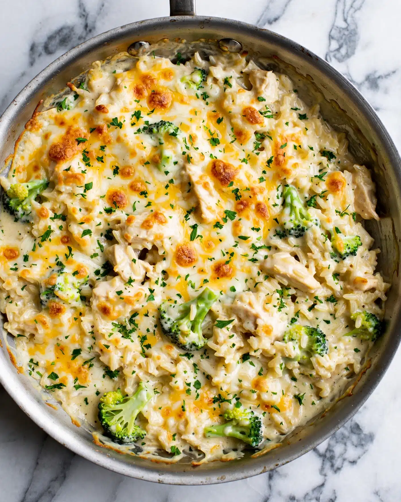A close-up top view of a silver skillet filled with a creamy rice dish that has a golden brown melted cheese layer on top mixed with white and yellow cheese. Inside, the dish contains small chunks of tender chicken, bright green broccoli florets, and creamy sauce blended with the rice, all topped with chopped fresh parsley sprinkled evenly across the surface. The skillet sits on a white marbled texture. photo taken with an iphone --ar 4:5 --v 7