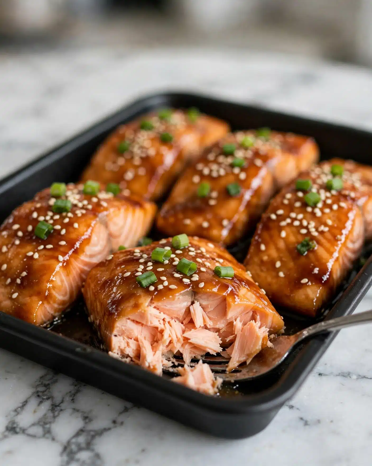The image shows several pieces of cooked salmon fillets placed close together in a black tray on a white marbled surface. Each salmon piece is covered with a shiny, sticky brown sauce and sprinkled with white sesame seeds and small pieces of bright green chopped scallions. The fillet in the front has a small section broken off revealing the soft, flaky, light pink inside of the salmon. A silver fork is partially visible under the broken piece, adding texture contrast. The background is softly blurred, focusing attention on the salmon. photo taken with an iphone --ar 4:5 --v 7