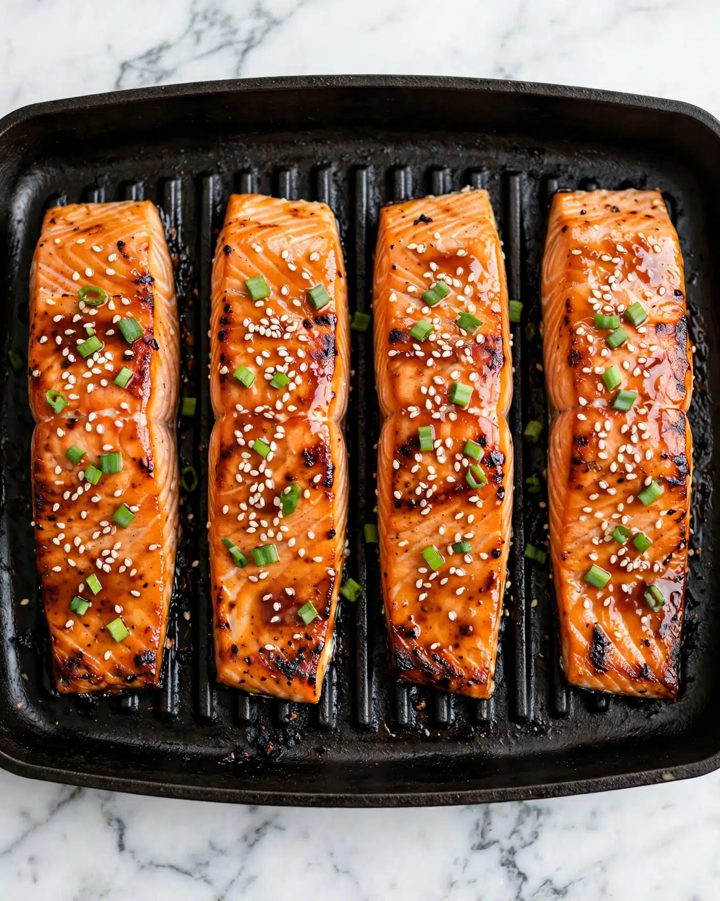 The image shows four grilled salmon fillets laid out side by side on a dark grill pan with clear grill marks. Each fillet is topped with melted glossy glaze, scattered white sesame seeds, and small chopped green onions which add a pop of color. The salmon has a rich orange-pink color with slightly charred edges giving it a cooked texture. The background is a white marbled surface. photo taken with an iphone --ar 4:5 --v 7