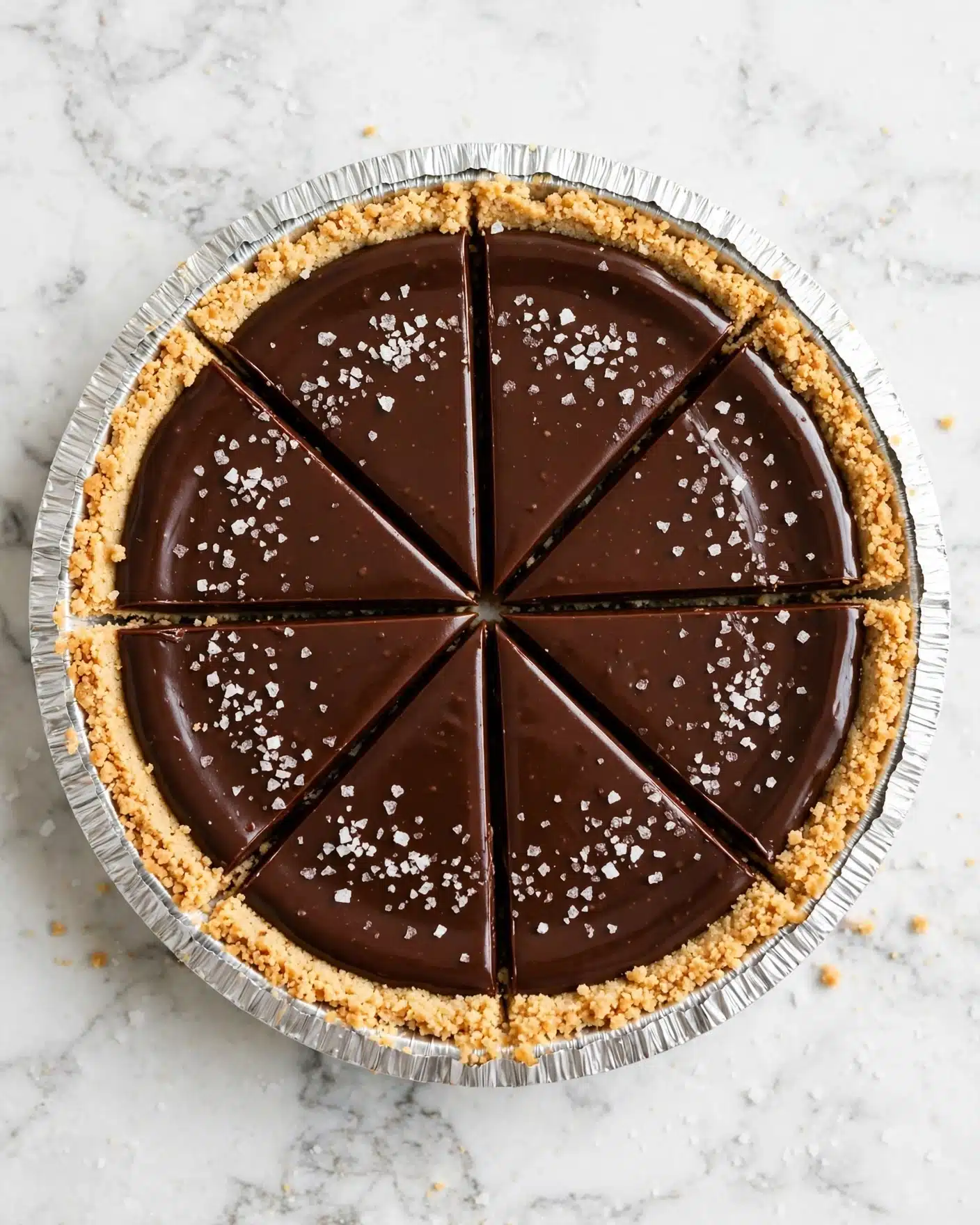 A round dessert with three clear layers is shown from above on a white marbled surface. The base layer is a crumbly, light golden crust forming the sides and bottom in a white foil pie pan. The middle layer is smooth and glossy dark brown chocolate filling that fills most of the pie and has cleanly sliced triangular segments. The top is sprinkled with coarse white sea salt flakes, adding texture to the shiny chocolate layer. Photo taken with an iphone --ar 4:5 --v 7