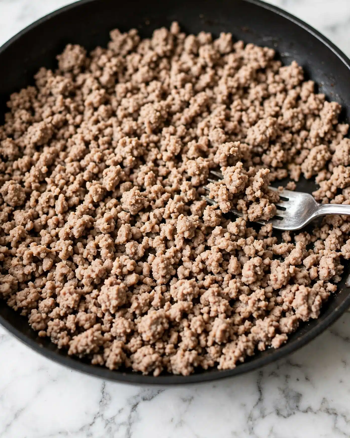 The image shows a close-up of a pan filled with cooked ground meat that is light brown in color. The texture of the meat is crumbly and evenly broken into small pieces. A silver fork rests on the right side of the pan, slightly lifting some of the meat. The pan has a dark, smooth edge and sits on a white marbled surface. Photo taken with an iphone --ar 4:5 --v 7