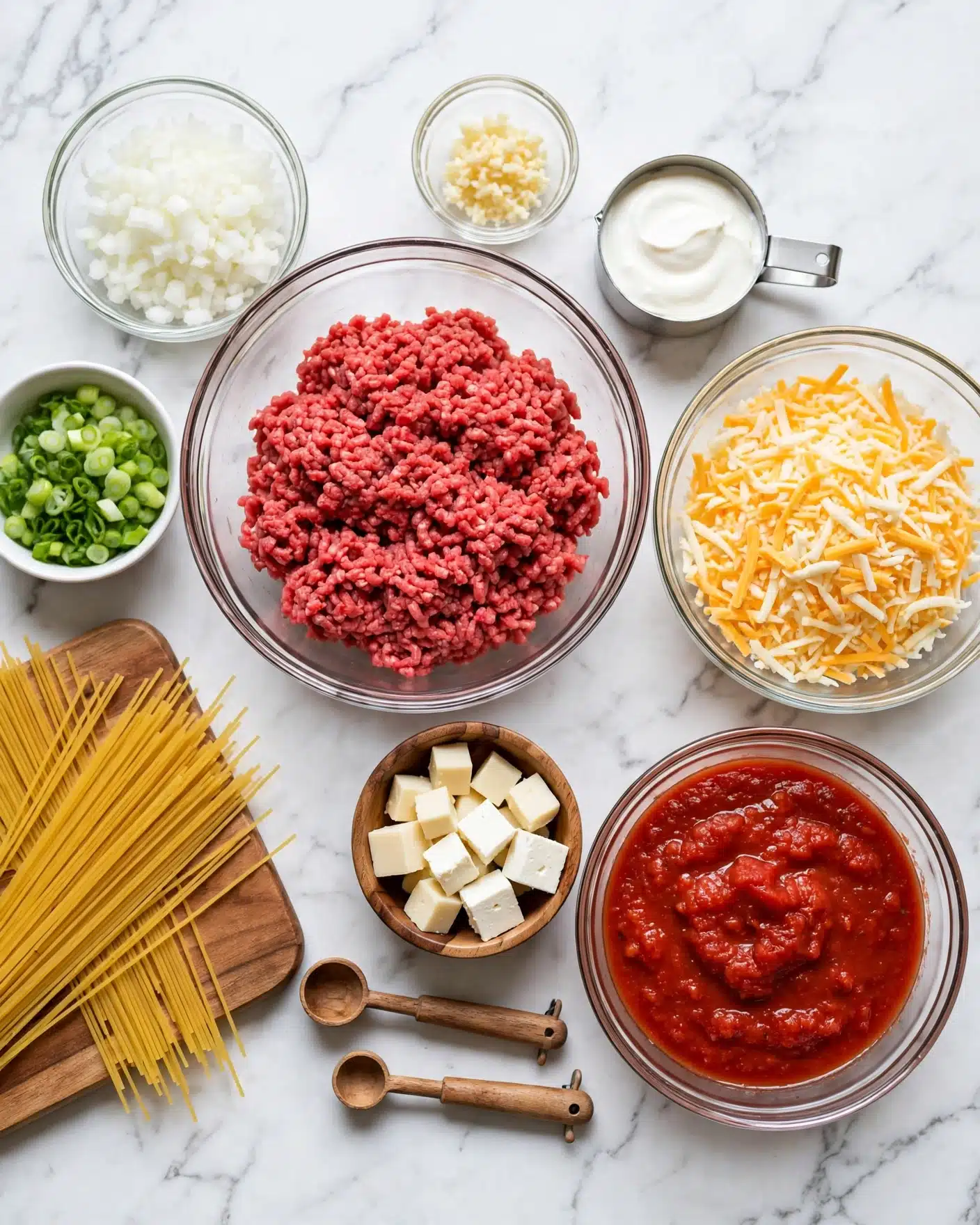 The image shows several clear glass bowls and a small wooden bowl arranged on a white marbled surface. The largest clear bowl in the center holds bright red raw ground meat with a soft, textured surface. To its right is a medium clear bowl filled with yellow and white shredded cheese, looking light and fluffy. Below that is another clear bowl containing thick red tomato sauce with a chunky texture. To the left of the sauce is a small wooden bowl with white cubed cheese. Above the ground meat bowl is a tiny clear bowl with minced garlic, pale yellow in color. Next to that is a metal measuring cup filled with smooth, white sour cream. Further left is a clear bowl with finely chopped, white chopped onions. Next to it is a small white bowl with finely sliced green onions, bright green and fresh. Below these bowls are two wooden spoons with wooden handles and small, round weighing scoops at the ends. A wooden board with dry uncooked spaghetti noodles rests on the lower left side. The whole setup looks clean and organized. Photo taken with an iphone --ar 4:5 --v 7