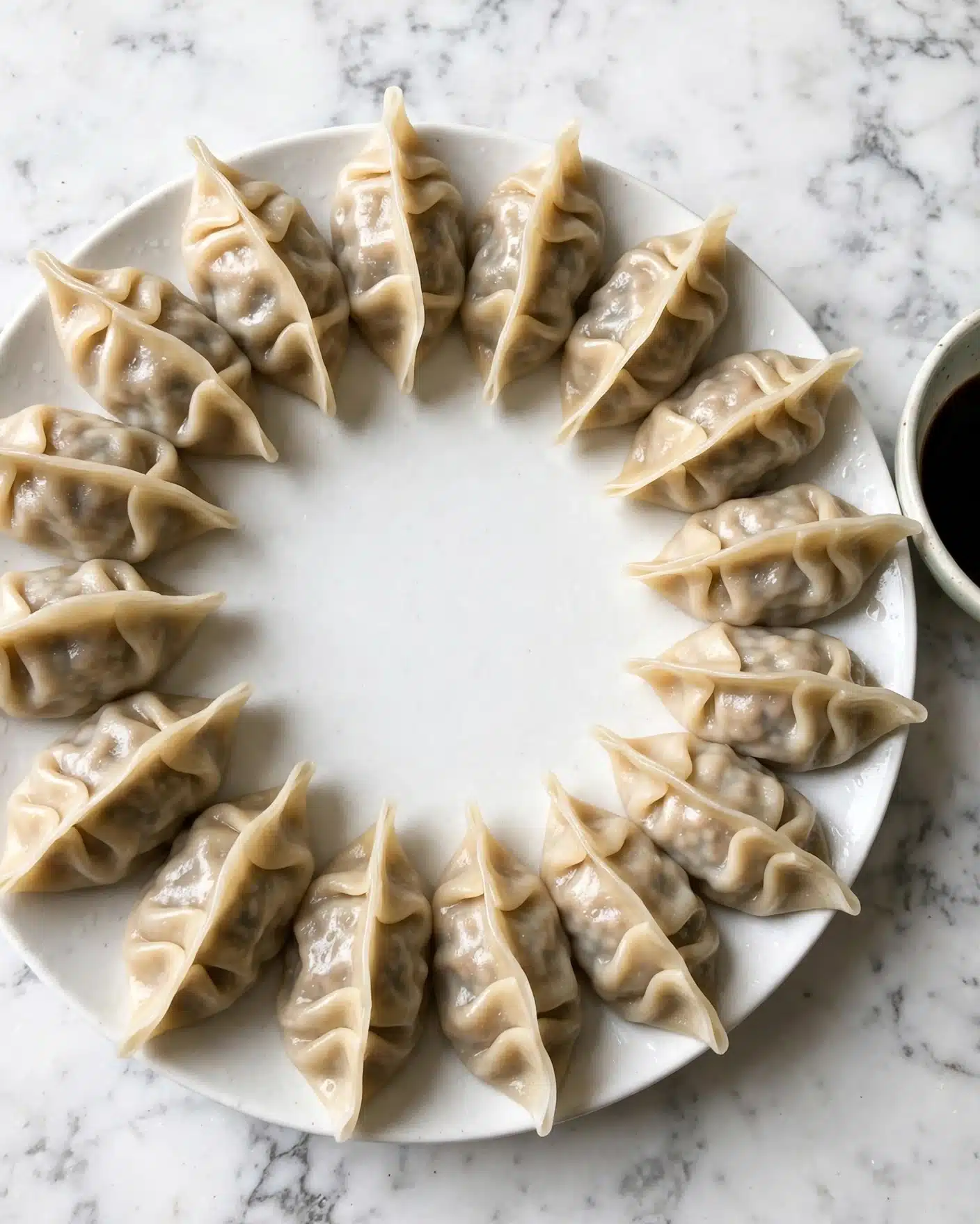 A white round plate holds sixteen steamed dumplings arranged in a circular pattern, each dumpling showcasing a light beige, soft, and slightly shiny translucent wrapper that reveals a subtle hint of a darker filling inside. The dumplings have gently folded edges creating a delicate texture on top, and they sit evenly spaced against a white marbled surface. A small bowl of dark soy sauce is partially visible to the right, adding contrast to the overall light tones. Photo taken with an iphone --ar 4:5 --v 7