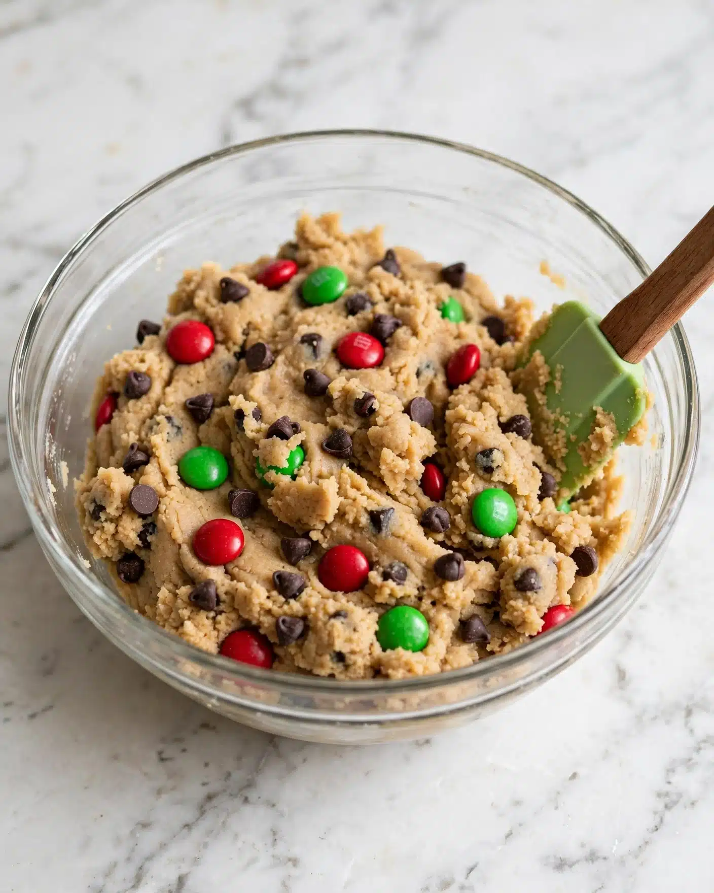 A clear glass bowl filled with light brown cookie dough mixed with small dark chocolate chips, bright red, and green candy-coated chocolates scattered throughout. The dough has a thick, soft texture with candy pieces slightly embedded on the surface. A spatula with a wooden handle and a light green rubber head is partially dipped into the dough on the right side of the bowl. The bowl is set on a white marbled surface. photo taken with an iphone --ar 4:5 --v 7