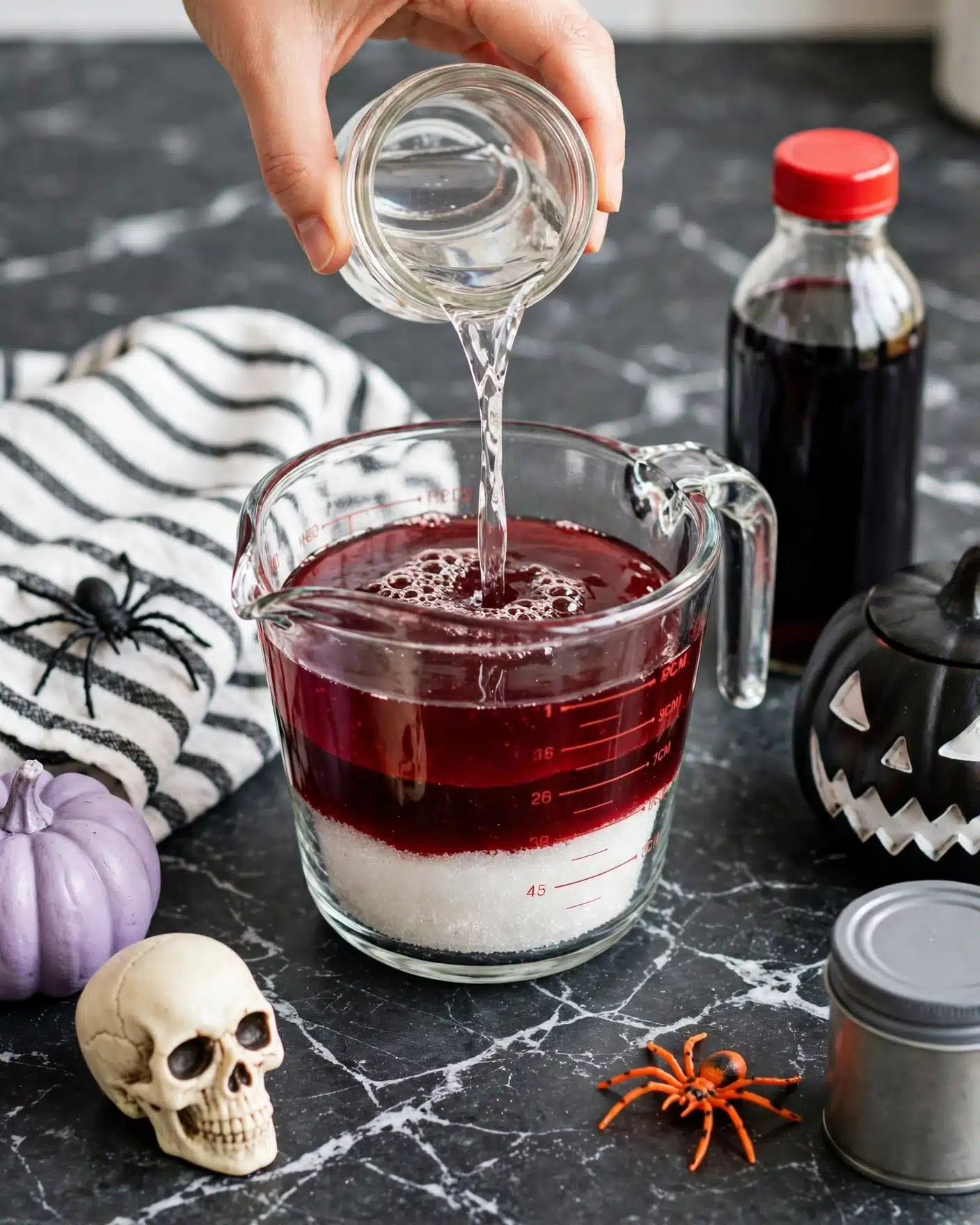 Vampire Kiss Drink Recipe 5 A clear glass measuring cup shows three layers inside: a bottom layer of bright white granulated sugar, a middle layer of thick, deep red syrup with a smooth texture, and an upper layer of clear liquid being poured in from a glass jar, held by a woman's hand. The cup sits on a dark countertop with a white marbled texture. Around it, there is a white and black striped cloth with a plastic black spider, a cream-colored skull with dark eye sockets, a small lavender pumpkin, a dark pumpkin-shaped container with a white jagged mouth design, a small orange plastic spider, and a metal can with a grey top. A bottle with a red cap and dark liquid inside stands near the cup. Photo taken with an iphone --ar 4:5 --v 7