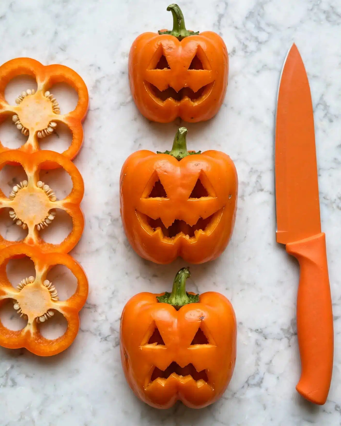 Jack-O'-Lantern Stuffed Peppers Recipe 5 The image shows three orange bell pepper tops on a white marbled surface, each carved with a different jack-o'-lantern face featuring triangle eyes and various mouth shapes, positioned vertically in the middle. To the left are three sliced orange bell pepper rings showing inner white seeds and walls, and to the right is an orange knife with an orange handle laying flat. The photo taken with an iphone --ar 4:5 --v 7