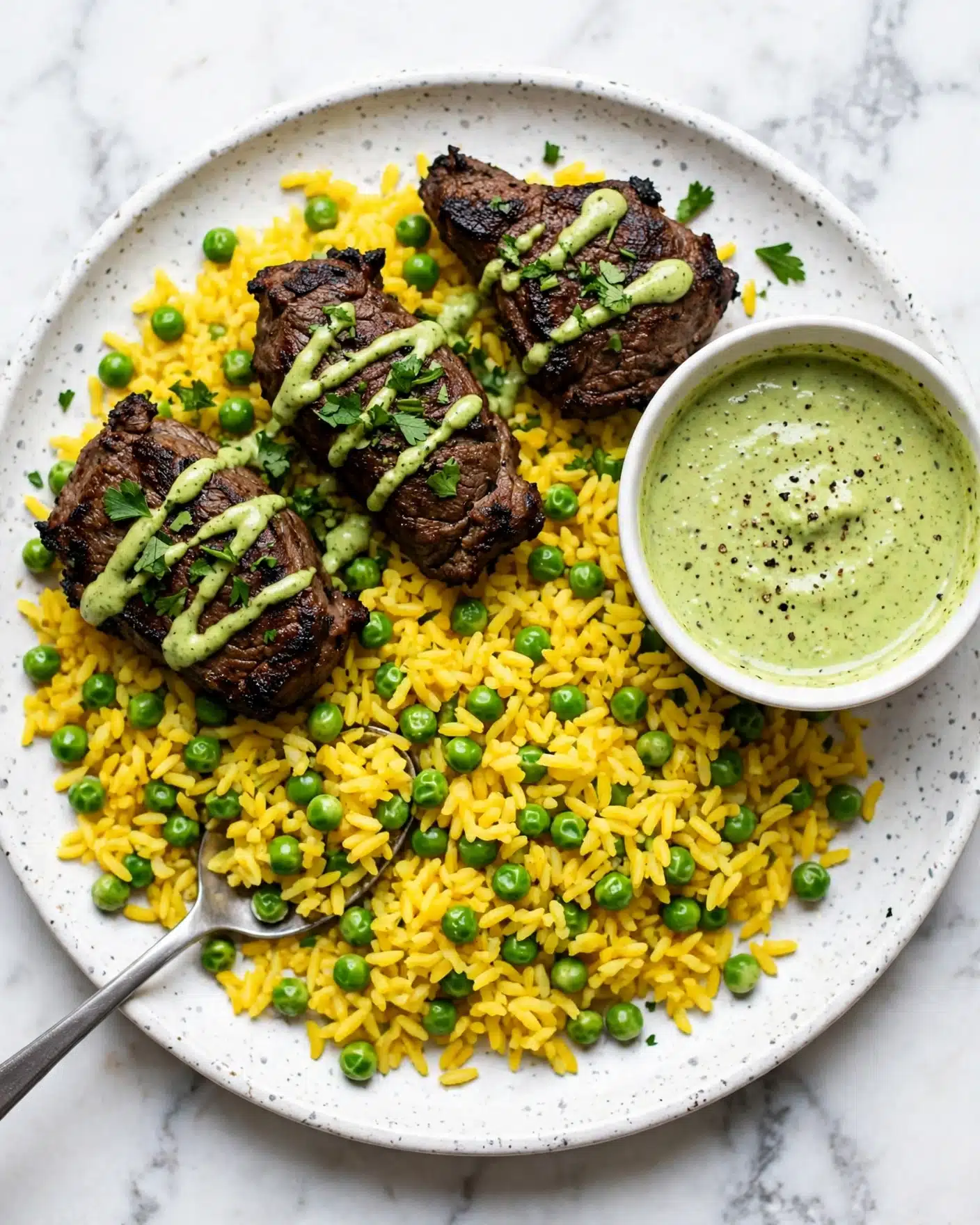 A white oval plate holds a bed of yellow rice mixed with bright green peas and herbs. On top are three grilled chicken pieces with dark, charred grill marks. The chicken is drizzled with a pale green sauce and sprinkled with fresh cilantro leaves. A silver spoon rests in the rice on the right side of the plate. In the bottom left corner, part of a small white bowl filled with the same pale green sauce is visible. The background has a white marbled texture. photo taken with an iphone --ar 4:5 --v 7