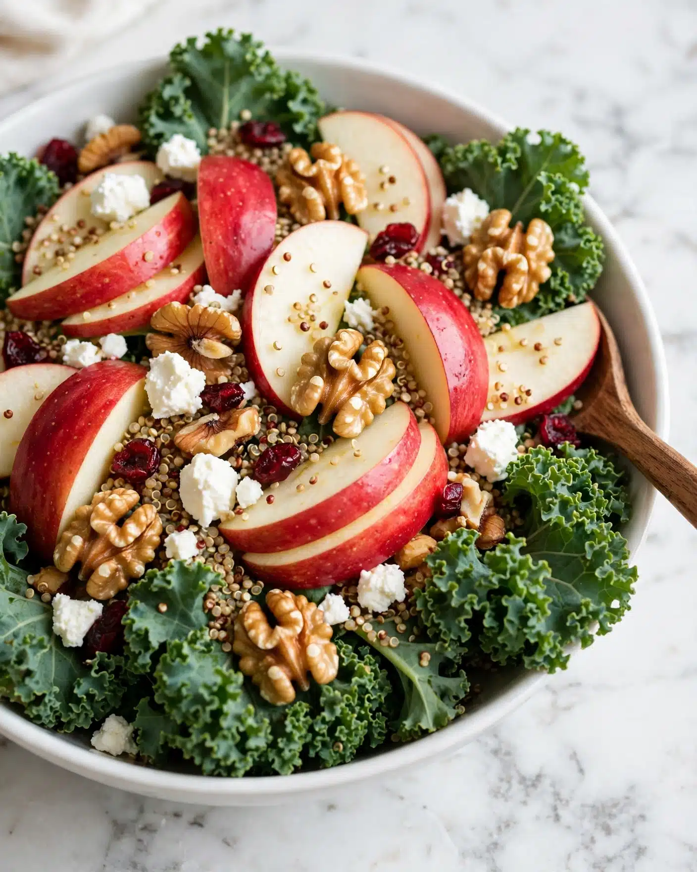 A white bowl full of fresh kale leaves with curly edges at the base, layered with thin slices of red-skinned apple showing crisp white inside scattered on top. Mixed within are small fluffy white cheese pieces and clusters of golden-brown walnut halves. Tiny red dried cranberries dot the salad, while tiny beige and brown quinoa grains are spread evenly across the layers, adding texture. The bowl rests on a white marbled surface, and a wooden spoon is partially visible on the right side. Photo taken with an iphone --ar 4:5 --v 7