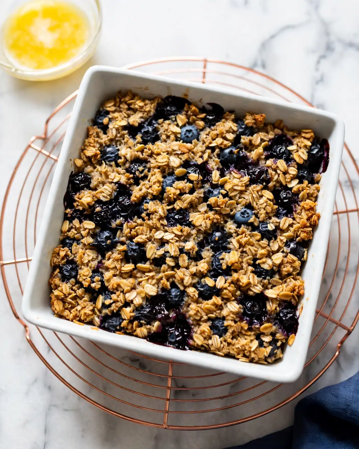 A white baking dish filled with a baked oatmeal layer, golden brown and textured with visible oats and chopped pecans scattered throughout, mixed with plump, deep blue and purple blueberries spread evenly on the surface. A spoon is scooping out a square section from the bottom right corner, revealing a moist and soft inner layer filled with more blueberries and oats. The dish is resting on a round copper metal trivet, all placed on a white marbled surface. photo taken with an iphone --ar 4:5 --v 7