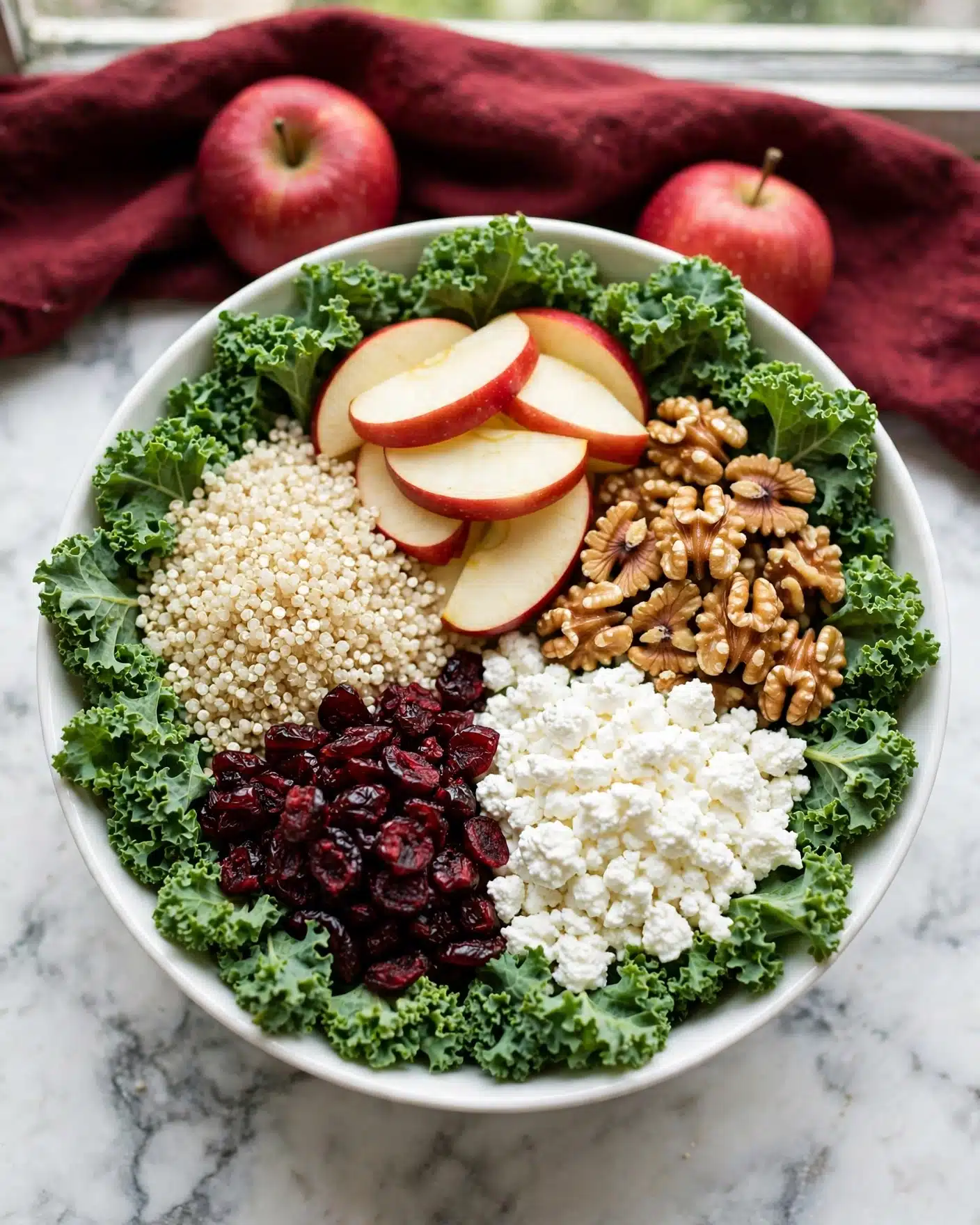A white bowl filled with five distinct layers of salad ingredients on a white marbled surface. The base layer is made of bright green curly kale leaves arranged around the bowl's edge. On top of the kale, there is a layer of small fluffy white quinoa grains on the left side, next to the quinoa is a pile of sliced red apple pieces with pale white flesh and red skin in the upper middle. To the right of the apples, there is a portion of golden-brown walnut halves, under them on the bottom right sits a pile of white cottage cheese crumbles, and next to the cheese on the bottom left is a heap of dark red dried cranberries. The scene includes red apples and a dark red cloth in the background. Photo taken with an iphone --ar 4:5 --v 7