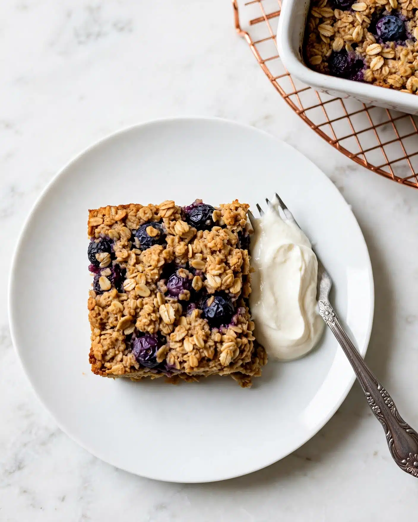 A square piece of baked oatmeal with visible whole blueberries and oats is placed near the center of a white plate, with a dollop of white cream on the side of the oatmeal, next to a silver fork with a decorated handle resting partly under the cream; the oatmeal has a rough, textured top with a mix of golden brown oats and purple blueberries, and the background is a white marbled surface with a corner of a white baking dish filled with similar oatmeal and blueberries visible at the top right corner, sitting on a round copper rack. photo taken with an iphone --ar 4:5 --v 7
