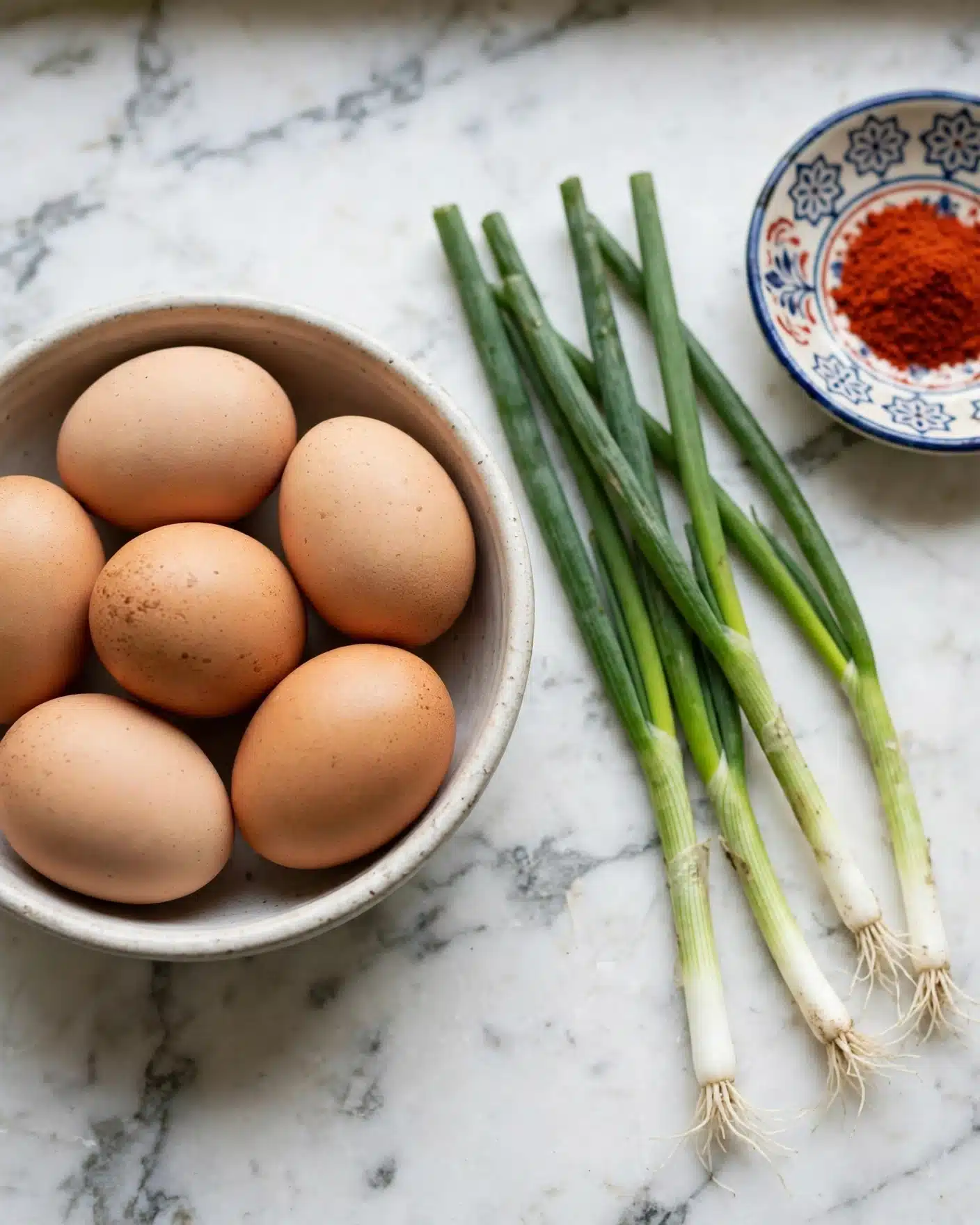Frittata Recipe, Plus 5 Variations Recipe 5 A round bowl filled with six brown eggs showing smooth, slightly speckled shells in shades of light brown. To the right of the bowl, five green onions lie on a white marbled surface, their long, slender stalks ranging from dark green at the top to white at the roots. Near the top right corner, a small white dish with colorful blue and red designs holds a bright red powder, adding a pop of color to the scene. The setting has soft, natural lighting, highlighting the textures and freshness of the ingredients. Photo taken with an iphone --ar 4:5 --v 7