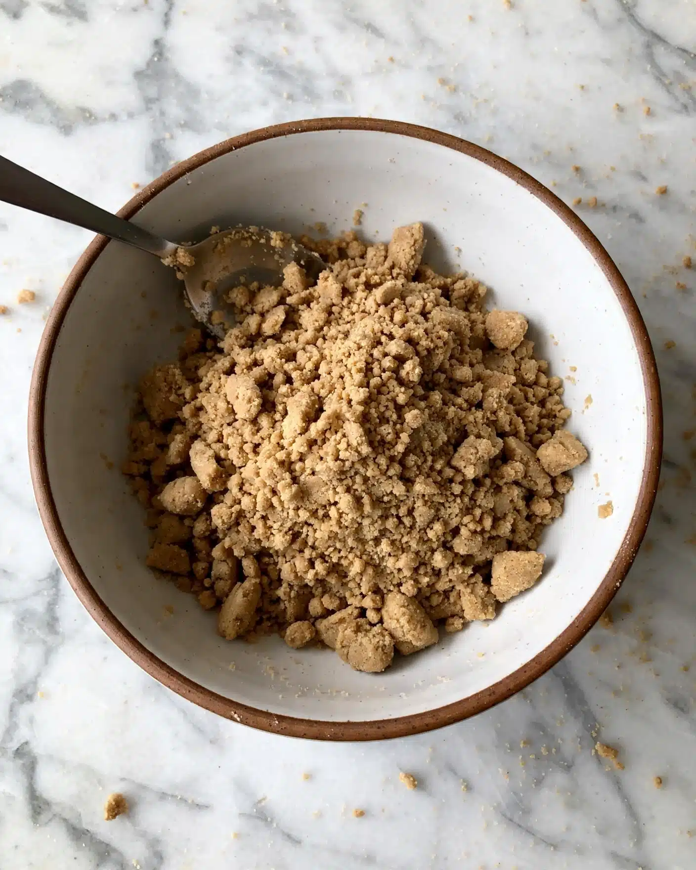A single white bowl with a brown rim holds a light brown, crumbly dough mixture, roughly piled in the center, showing a coarse texture with small clumps. A metal spoon rests inside the bowl on the upper left side, with some dough stuck to it. The bowl is placed on a white marbled surface with small scattered crumbs around. photo taken with an iphone --ar 4:5 --v 7