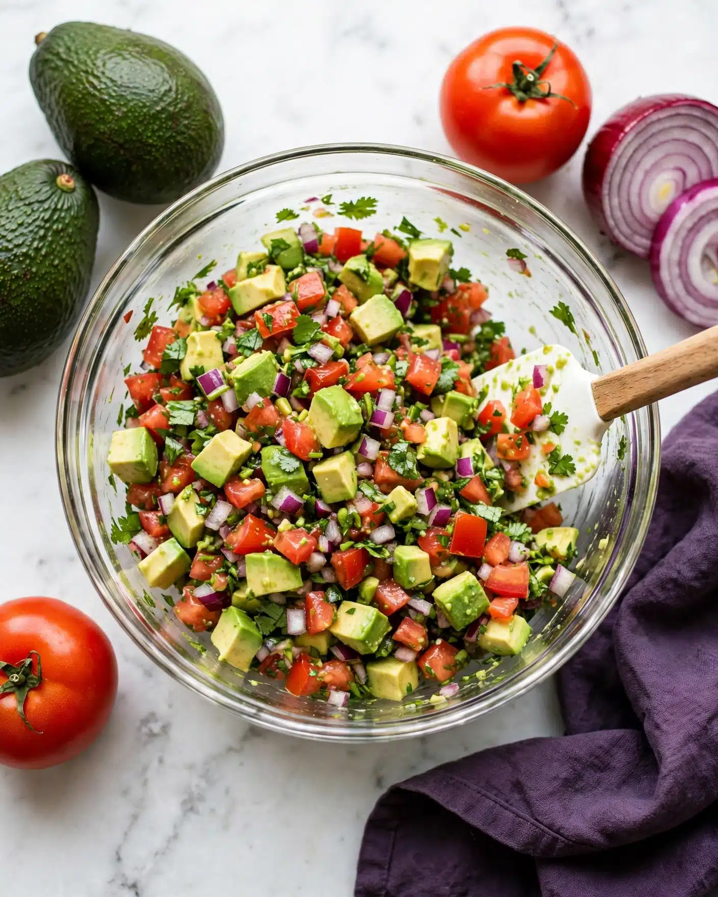 A clear glass bowl filled with a colorful mix of diced avocado, red tomatoes, red onion, and chopped green herbs, all finely mixed together showing fresh and vibrant colors. The bowl sits on a white marbled surface with fresh avocados, whole tomatoes, and a sliced red onion placed around it. A white spatula with a wooden handle is resting inside the bowl on the right side, partially covered by the mix. The texture looks fresh and slightly chunky, with the diced ingredients clearly seen. A dark purple cloth lies next to the bowl on the surface. Photo taken with an iphone --ar 4:5 --v 7